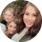Smiling woman with long brown hair wearing a white sleeveless top and white pants sitting on a beige tufted chair.