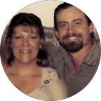 Smiling woman with long hair wearing a straw hat and necklace standing in a grassy field under a cloudy sky.