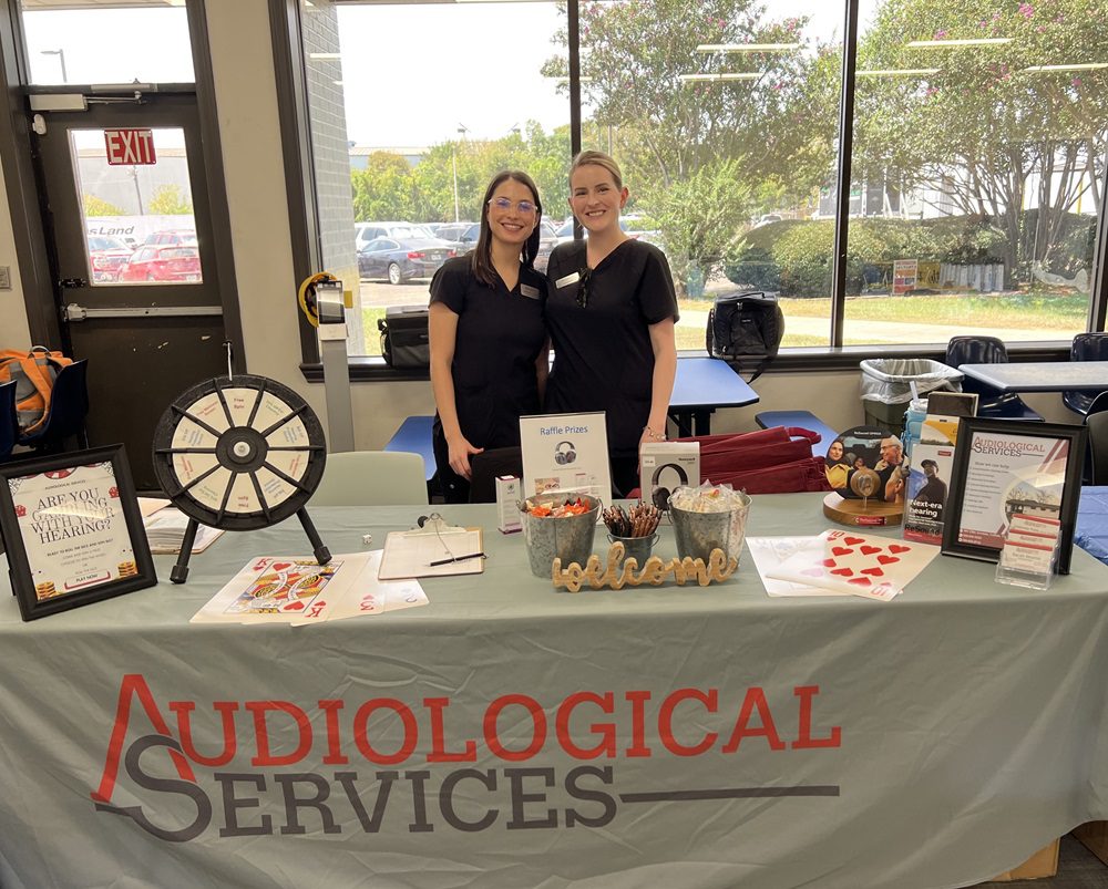 Person standing behind a booth table covered with informational pamphlets, hearing aids, and a rifle prop, with a black backdrop and a sign reading 'All About Hearing'.