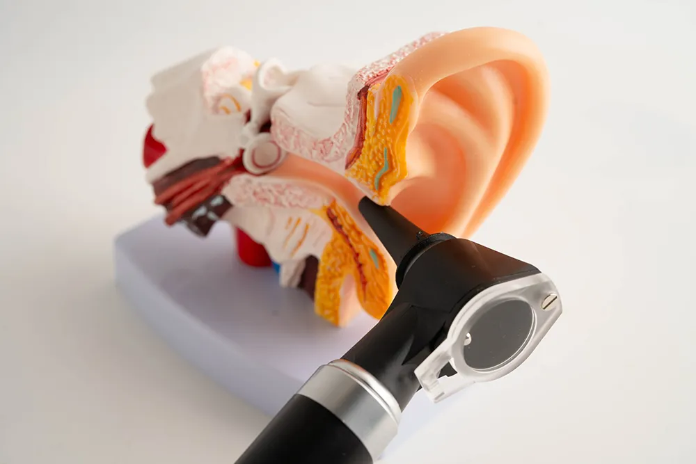 Female audiologist smiling and sitting at a desk with an otoscope and hearing aid on it, a computer with audiology software in the background.