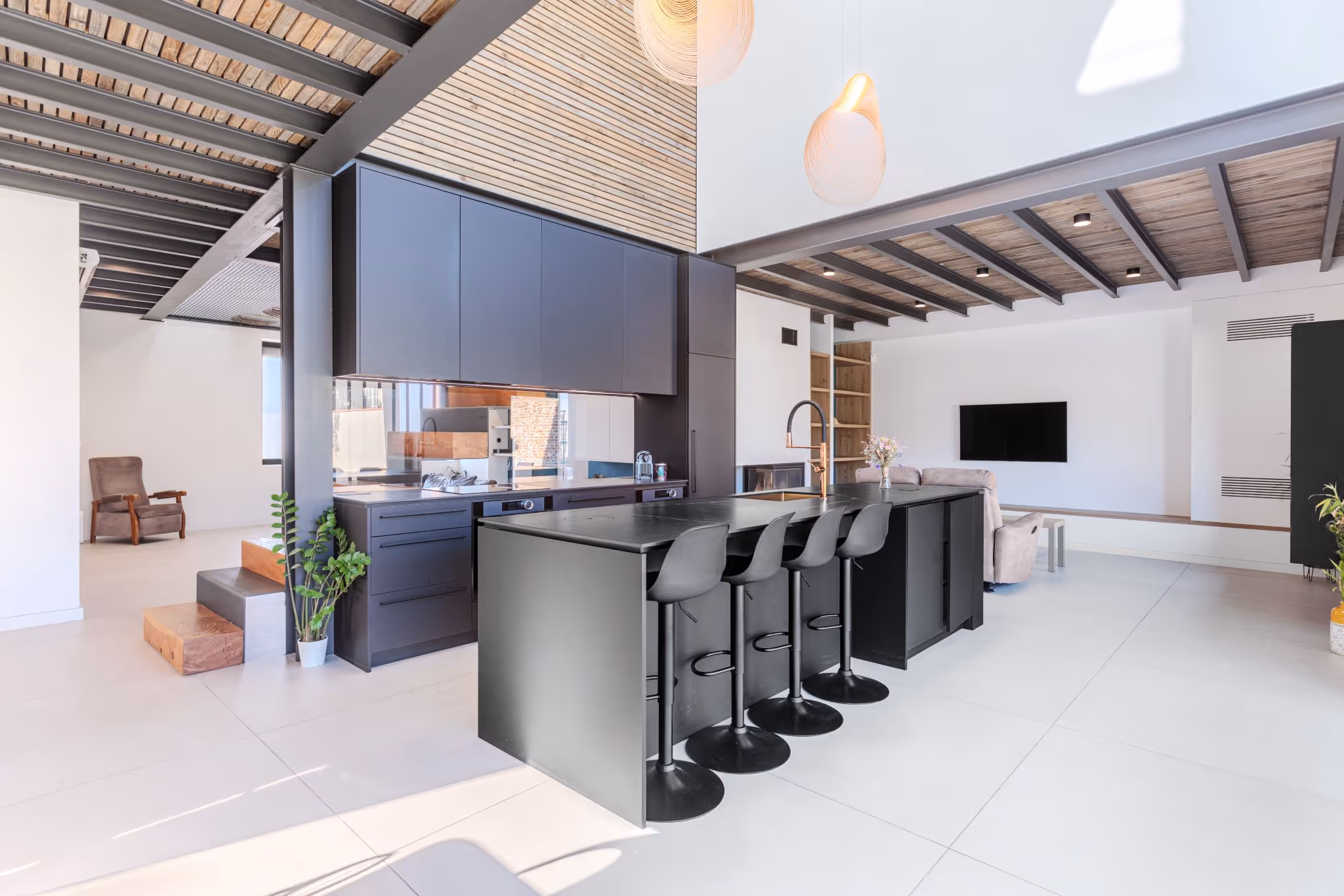 Cuisine moderne ouverte avec îlot central noir, quatre tabourets, plafond en bois avec poutres apparentes et salle de séjour avec canapé et téléviseur mural.