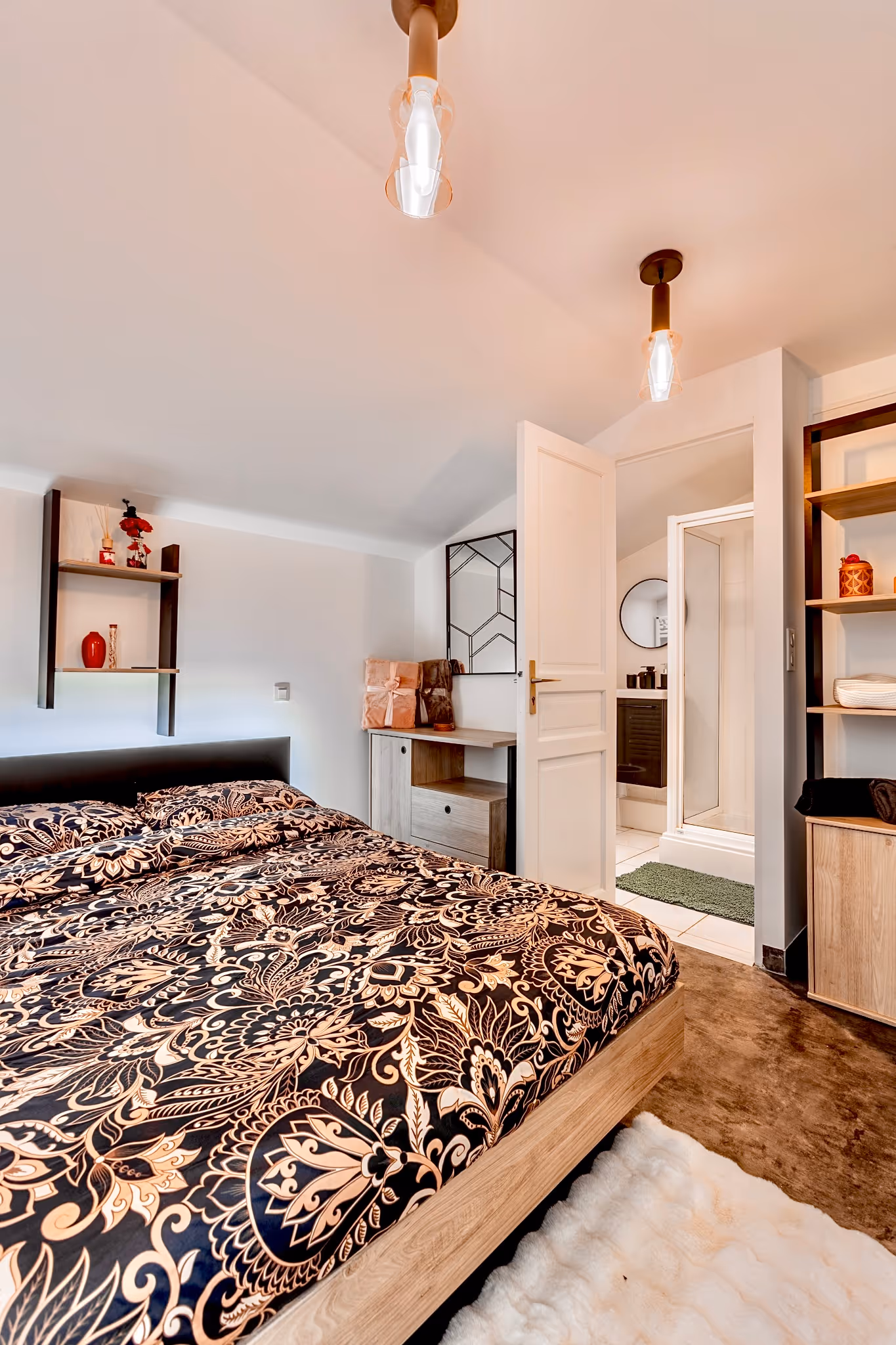Chambre moderne avec lit double à couette noire et beige à motifs floraux, meubles en bois clair, et salle de bain visible par une porte ouverte.