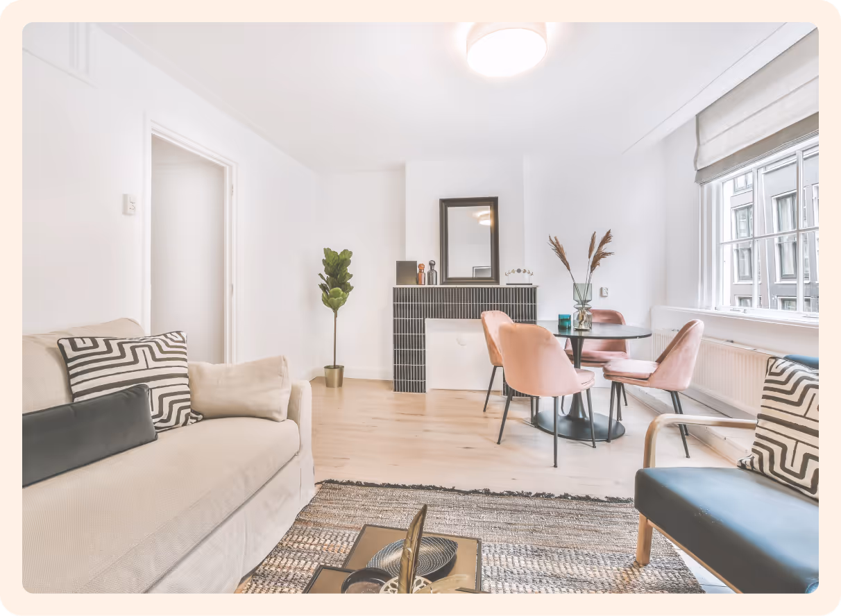 Salon lumineux avec canapé beige, fauteuil noir, table ronde avec chaises roses et grande fenêtre.