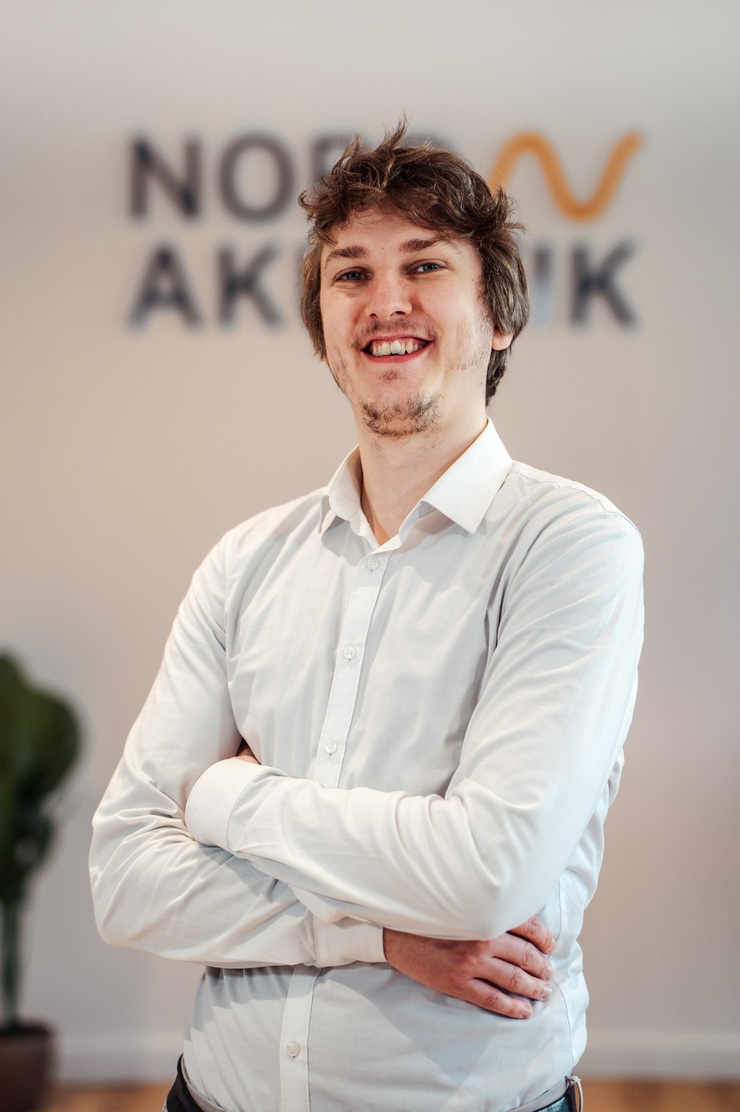 Ein lächelnder Mann in blauem Pullover und hellen Hosen läuft über einen gepflasterten Platz vor einem gelben Gebäude mit verzierten Fenstern.