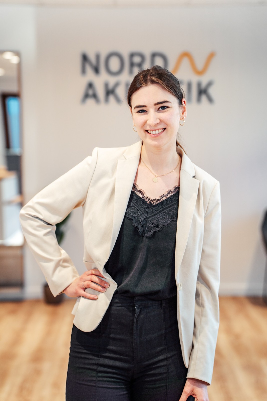 Junge Frau in beigem Blazer und schwarzer Spitzenbluse lächelt vor einer Wand mit dem Schriftzug 'Nord Akustik'.