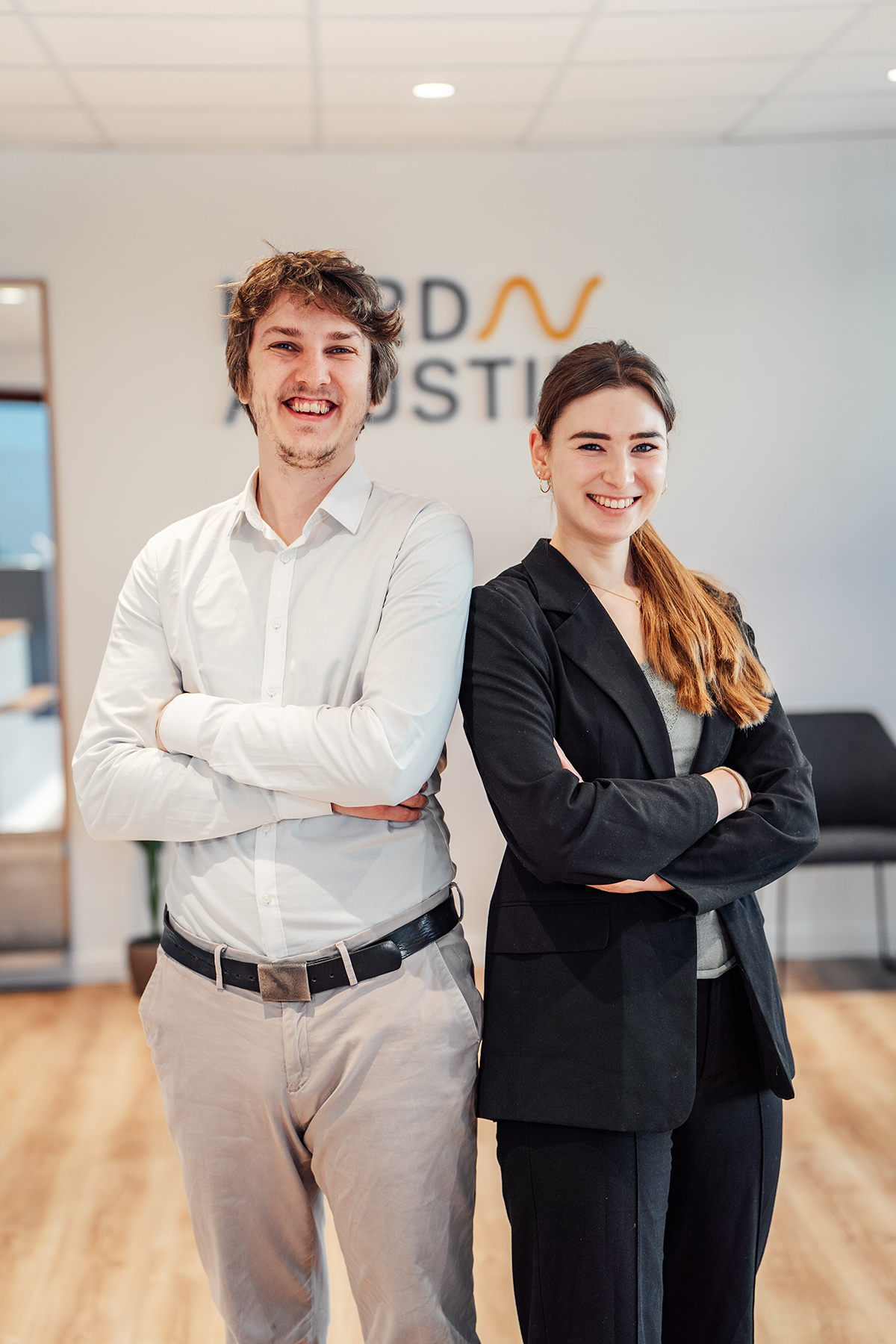 Junger Mann mit blauem Hemd und beiger Hose steht lächelnd vor einem gelben Gebäude.