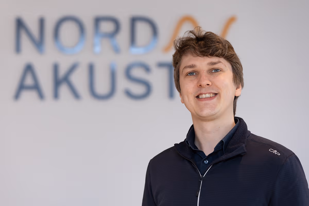 Ein lächelnder Mann in blauem Pullover und hellen Hosen läuft über einen gepflasterten Platz vor einem gelben Gebäude mit verzierten Fenstern.