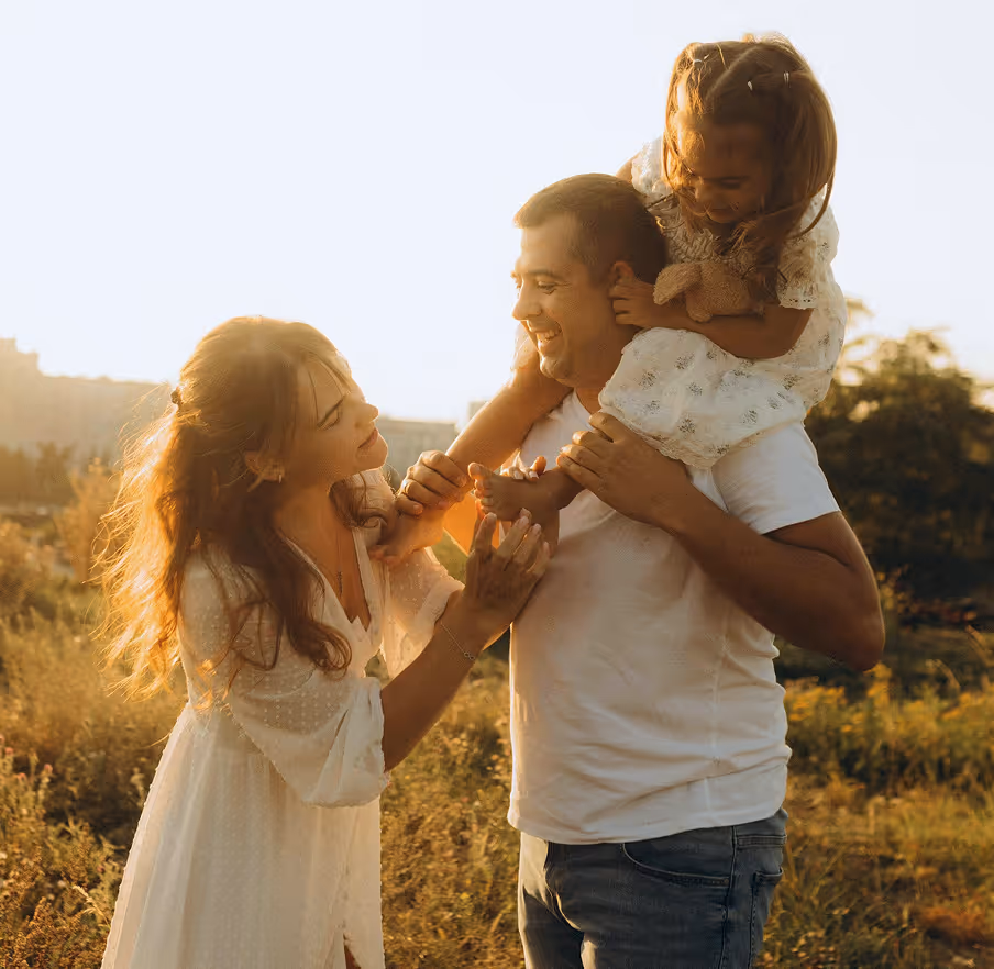 Smiling man carrying a young girl on his shoulders while a woman in a white dress playfully touches the girl's foot outdoors at sunset.