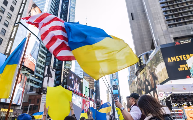Crowd in an urban area waving Ukrainian flags and an American flag among tall buildings and electronic billboards.