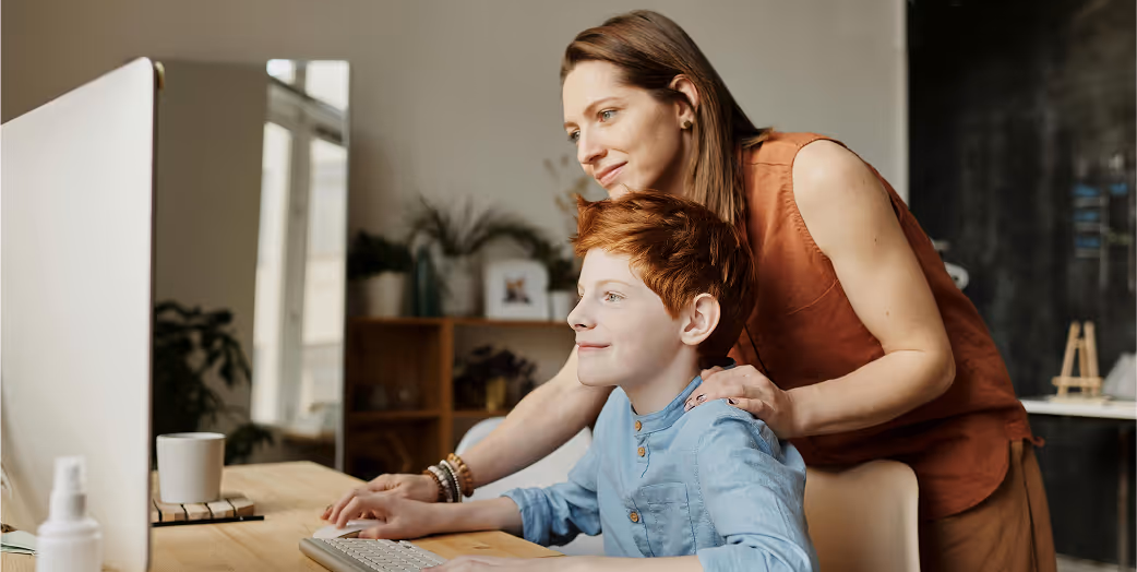 Woman guiding a young boy using a computer at a wooden desk in a cozy room.