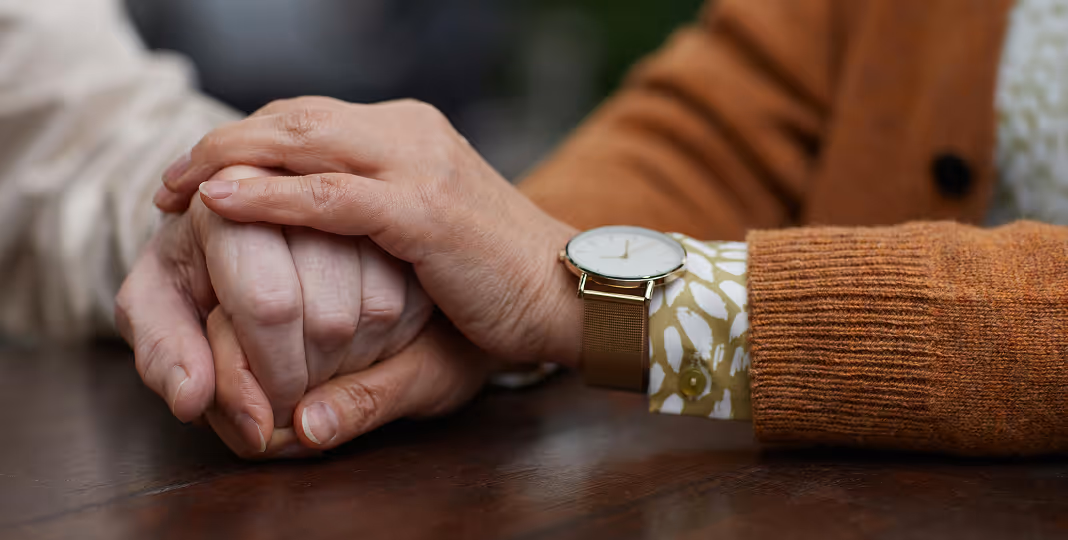 Close-up of two people holding hands on a wooden table, one wearing an orange sweater and wristwatch.