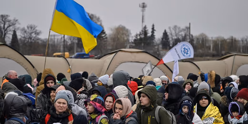 Crowd of people outdoors in winter clothing with a large Ukrainian flag and medical aid flags in the background.