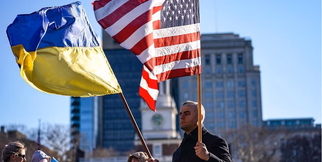 Man holding Ukrainian and American flags in a city square with buildings in the background.