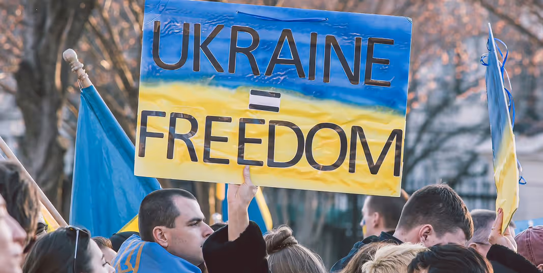 Crowd holding Ukrainian flags and a sign reading 'UKRAINE = FREEDOM' in blue and yellow colors.