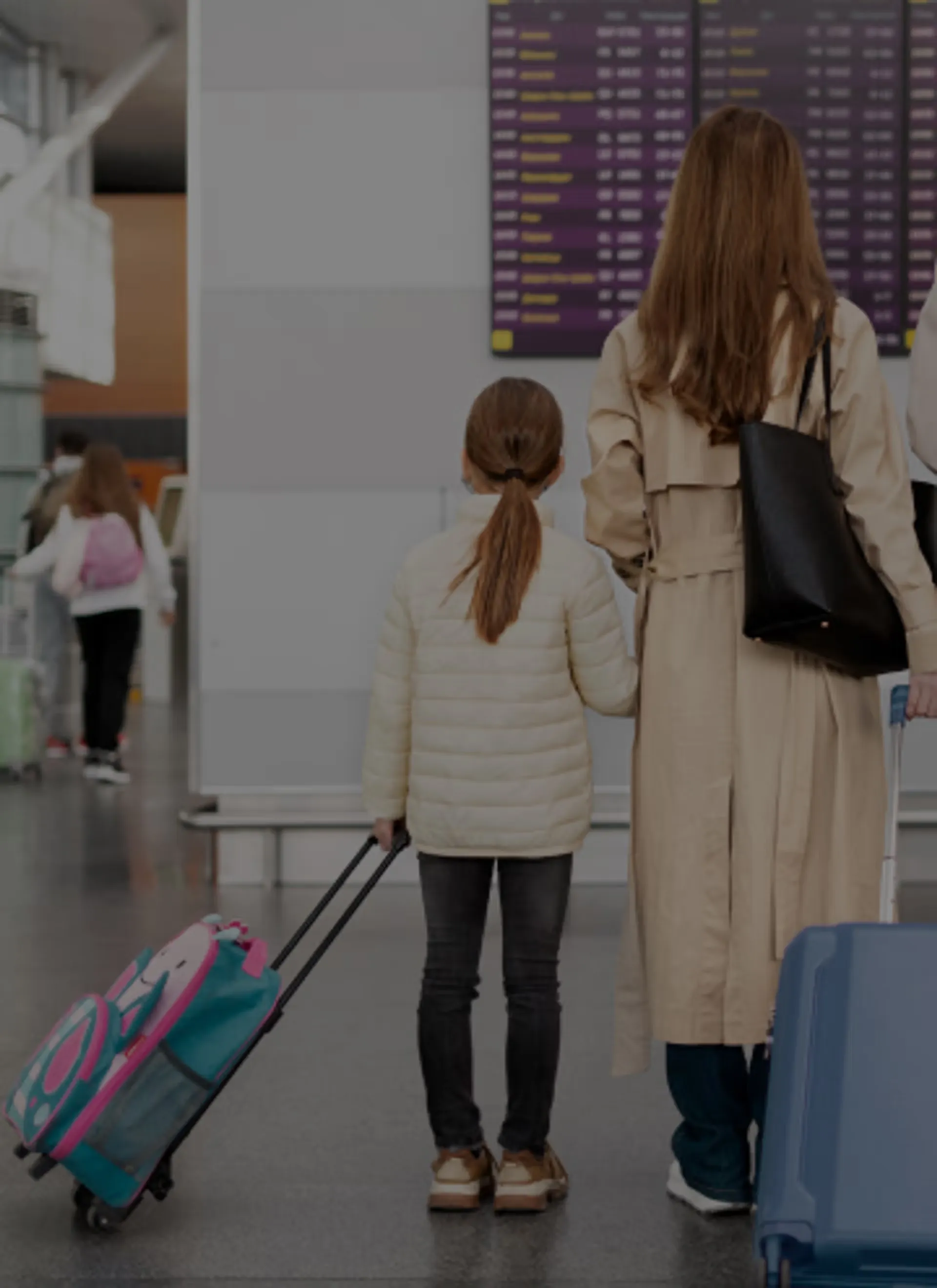 Woman and child holding hands while pulling suitcases, looking at a flight information board in an airport.