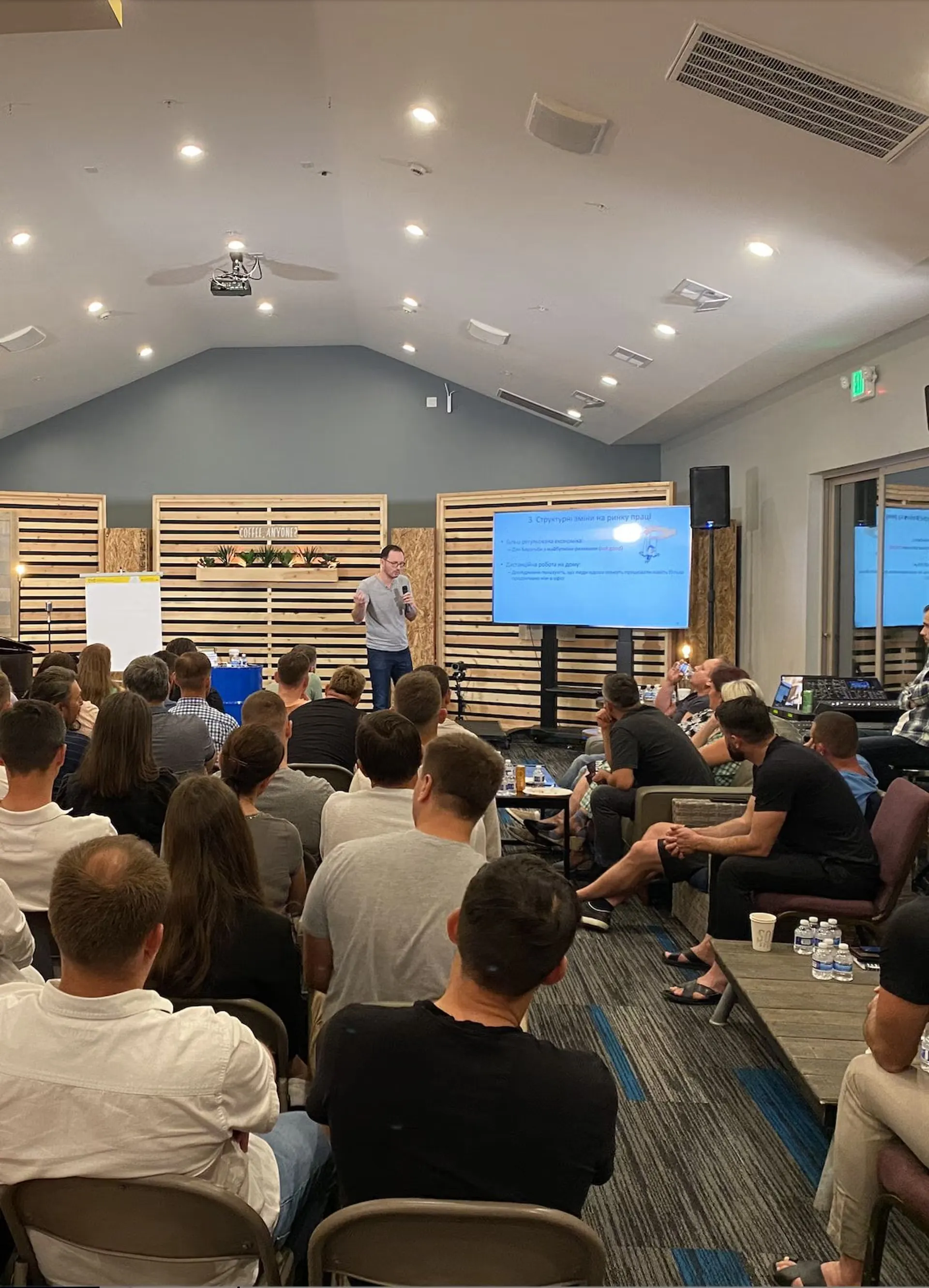 A man giving a presentation to a seated audience in a modern conference room with a screen displaying slides.