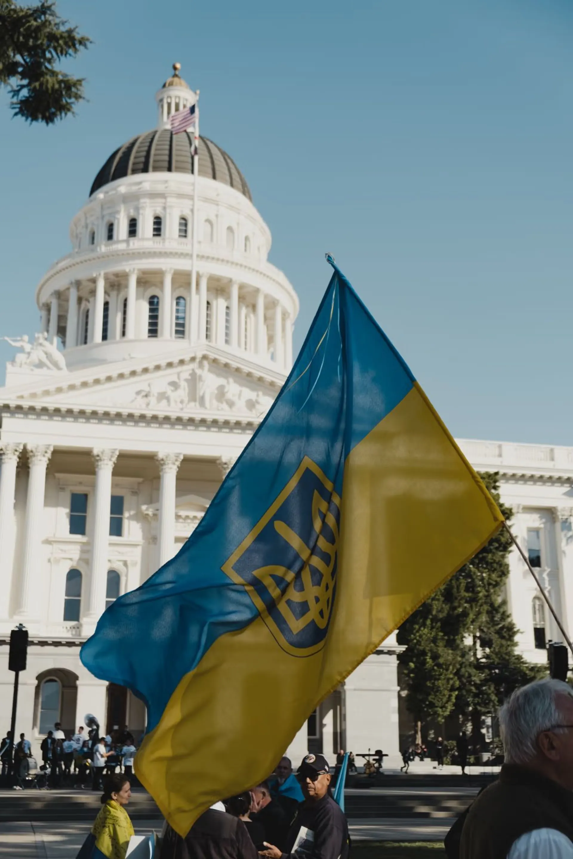 People holding a large blue and yellow Ukrainian flag with the state emblem in front of a white domed government building.