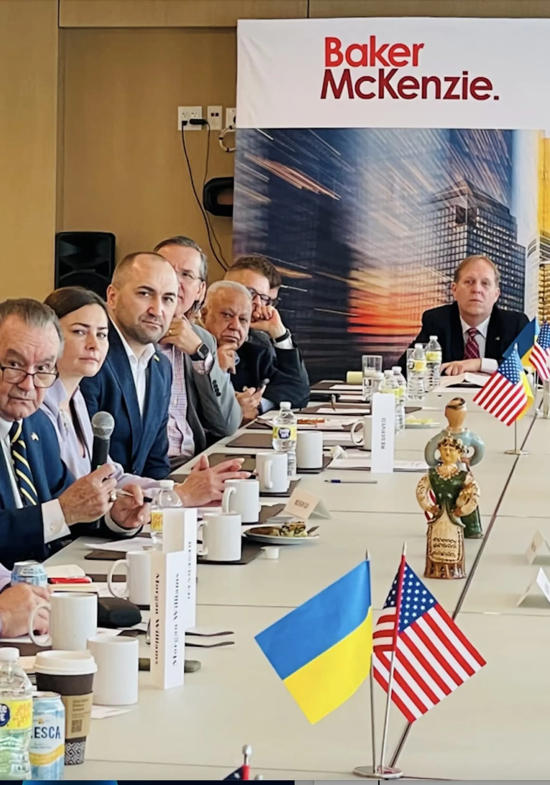 Group of people seated at a conference table with Ukrainian and American flags and a Baker McKenzie banner in the background.