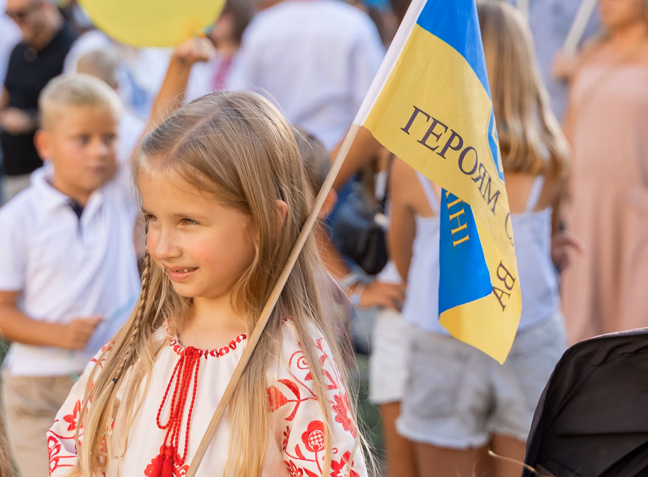 Young girl wearing a traditional embroidered shirt holding a blue and yellow flag with Cyrillic text, with people blurred in the background.