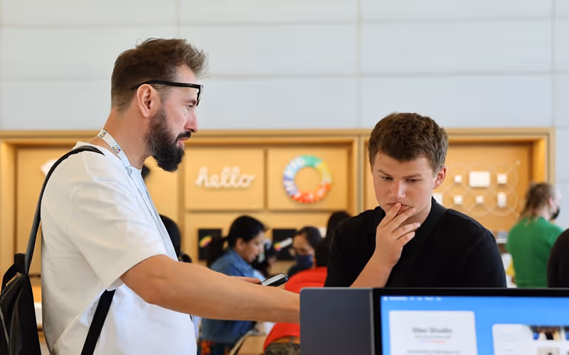 Man with beard and glasses instructing a young man at a computer in a casual tech store setting.