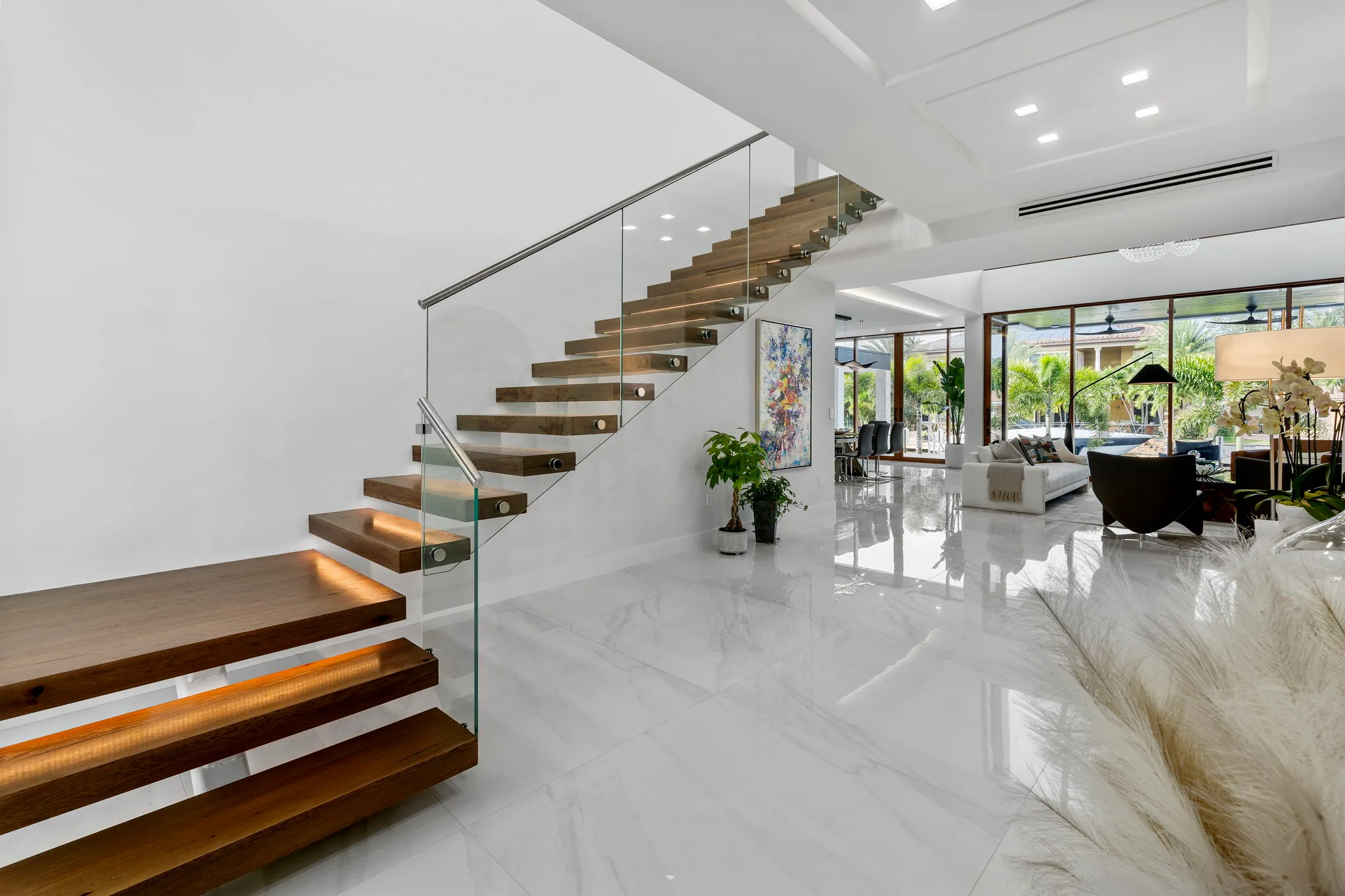 Modern interior with floating wooden stairs featuring glass railing and ambient lighting, leading to a bright living area with large windows.