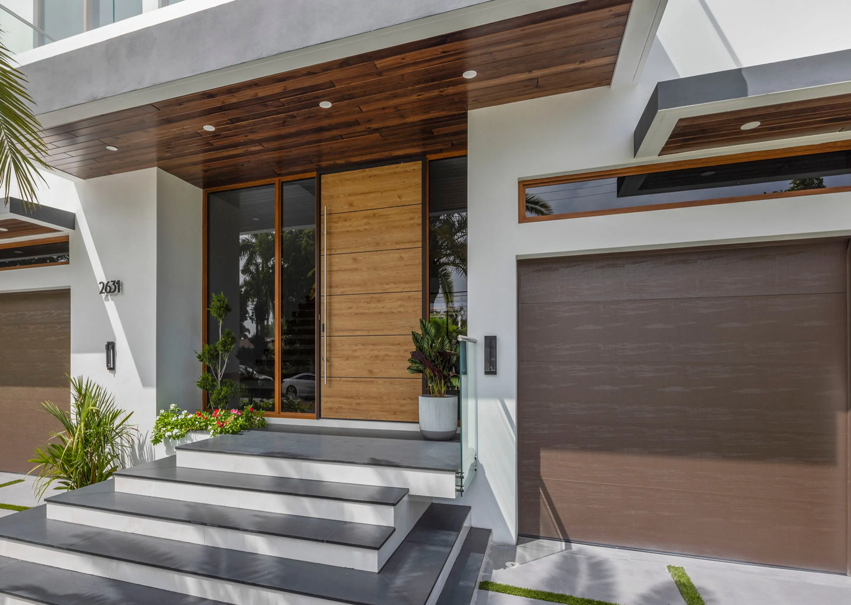 Modern house entrance with wide wooden door, glass side panel, gray steps, and brown garage door.