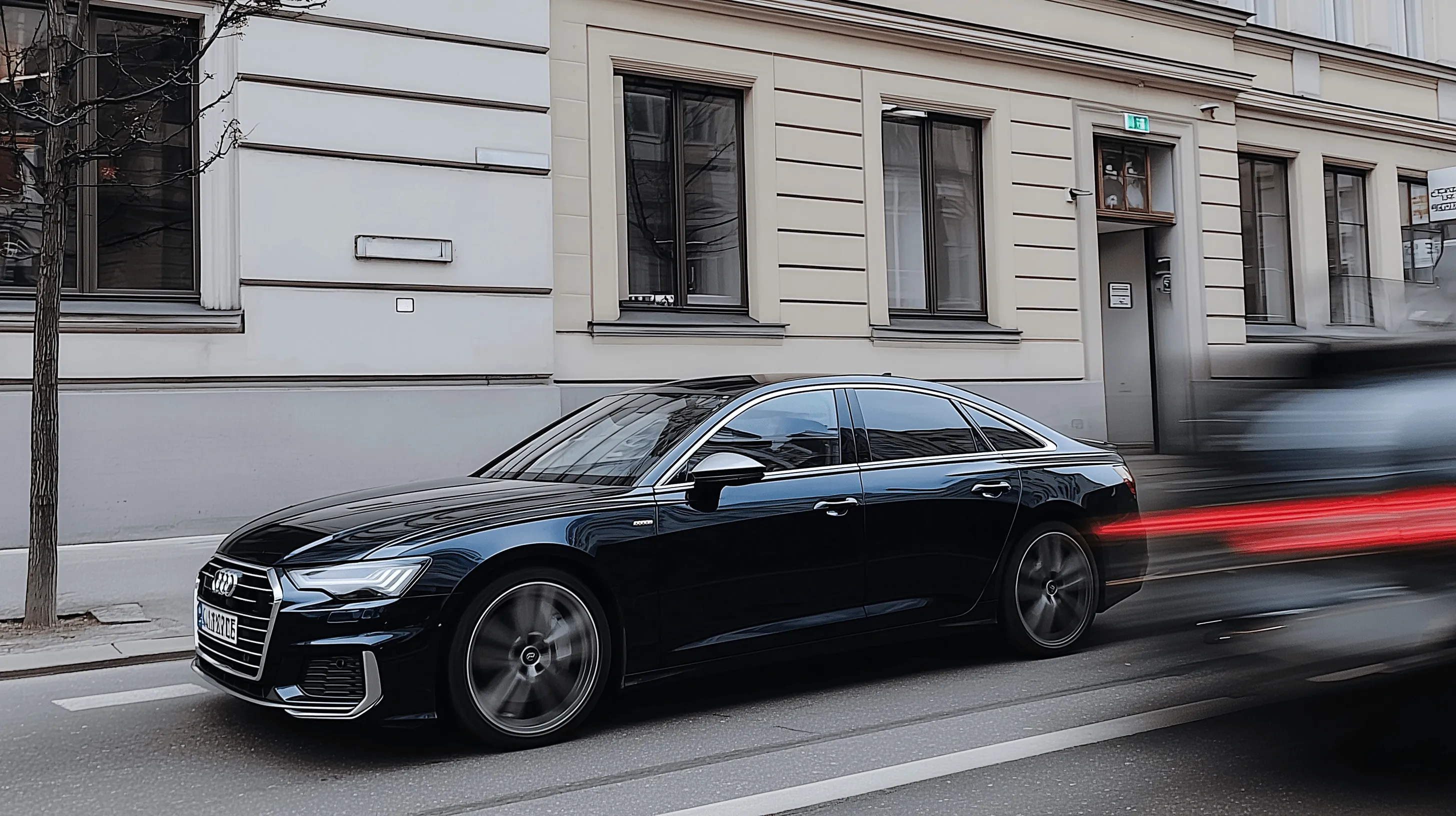 Black Audi sedan driving on a city street with building windows in the background and another vehicle passing quickly causing red light streaks.