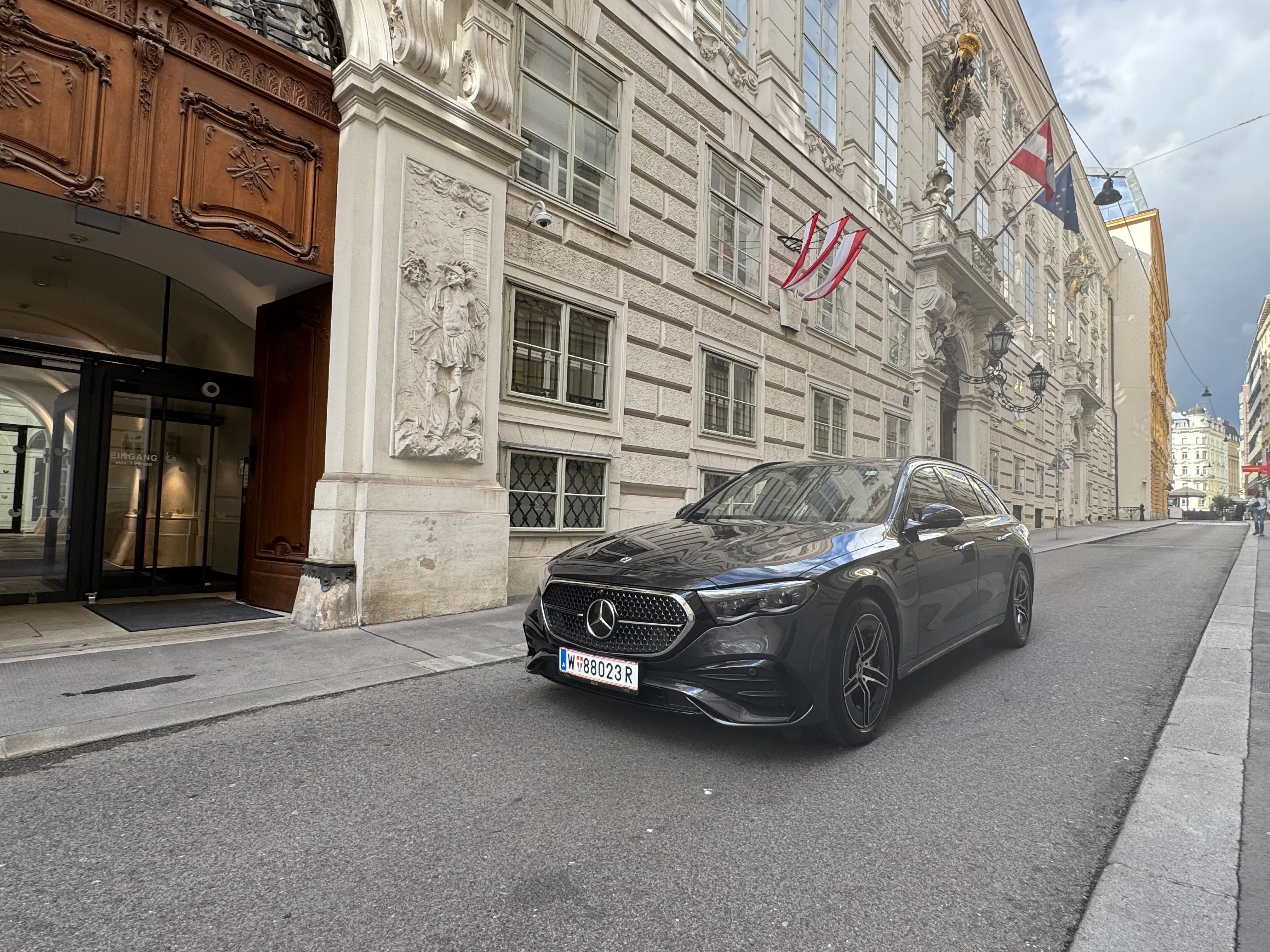 Black Mercedes-Benz car parked on a narrow street in front of a historic building with ornate stone carvings and several flags.