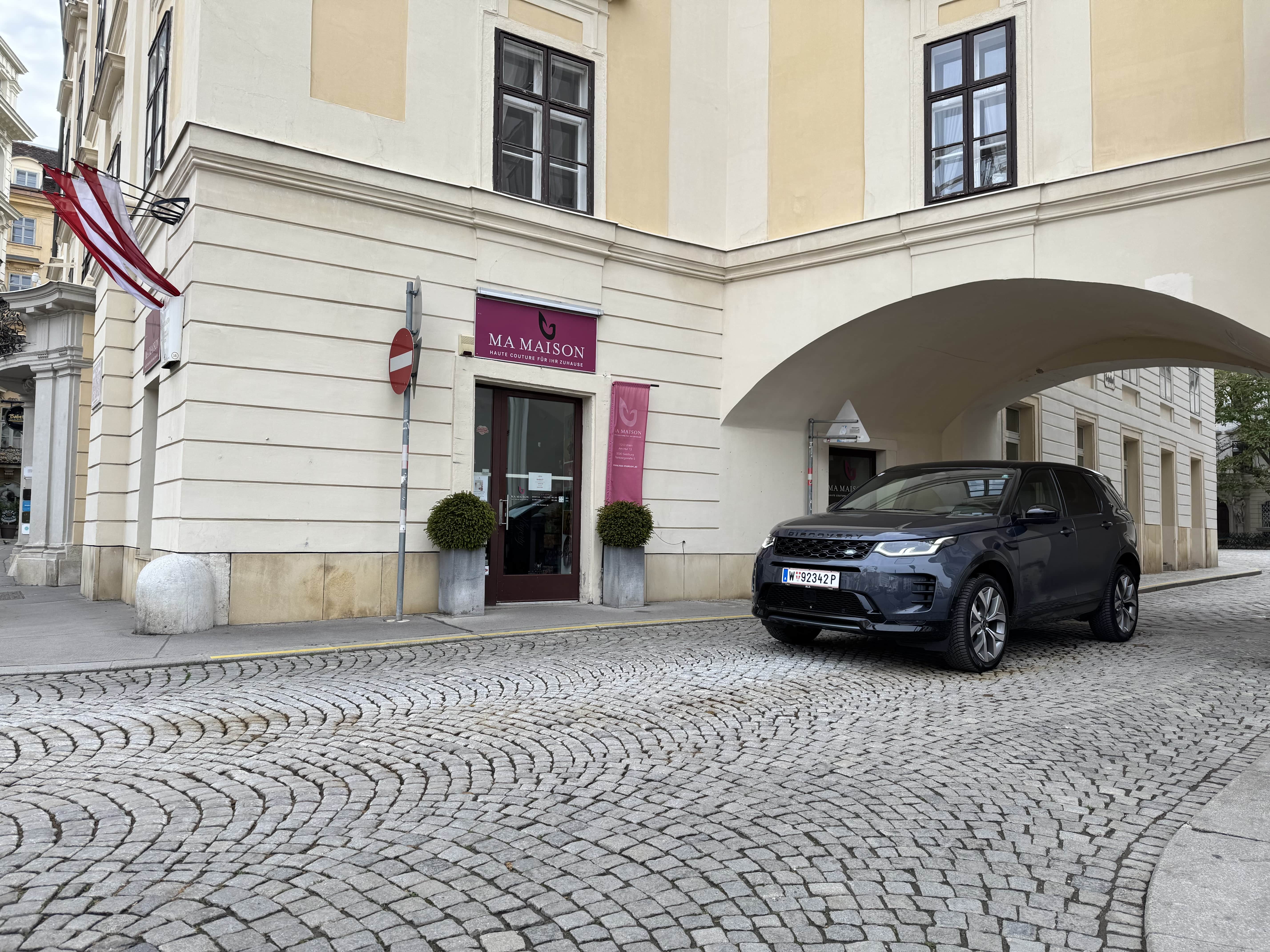 Dark blue Land Rover Discovery parked on a curved cobblestone street next to a beige building with a maroon sign reading 'MA MAISON'.