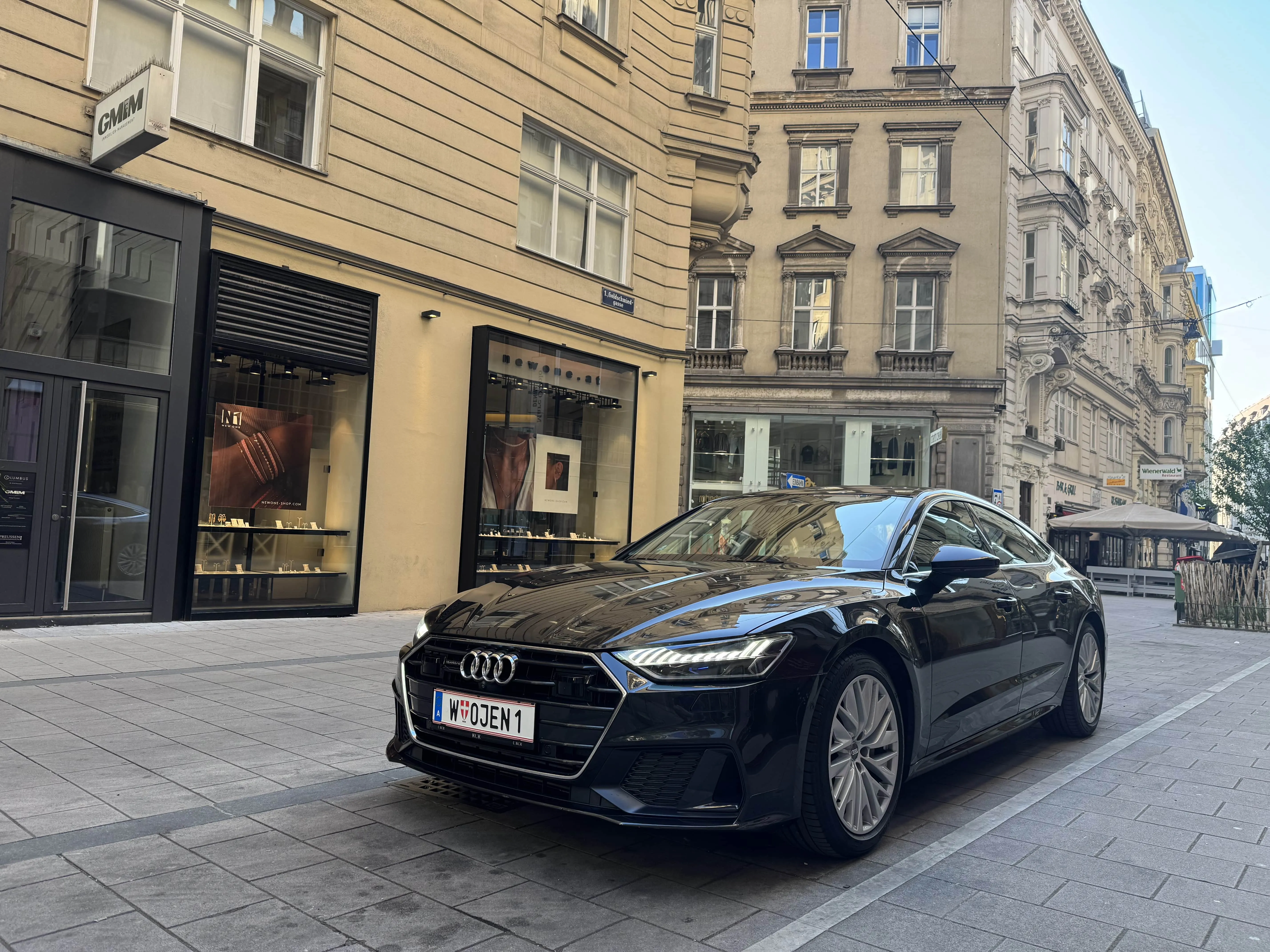 Black Audi sedan parked on a cobblestone city street in front of beige European-style buildings.