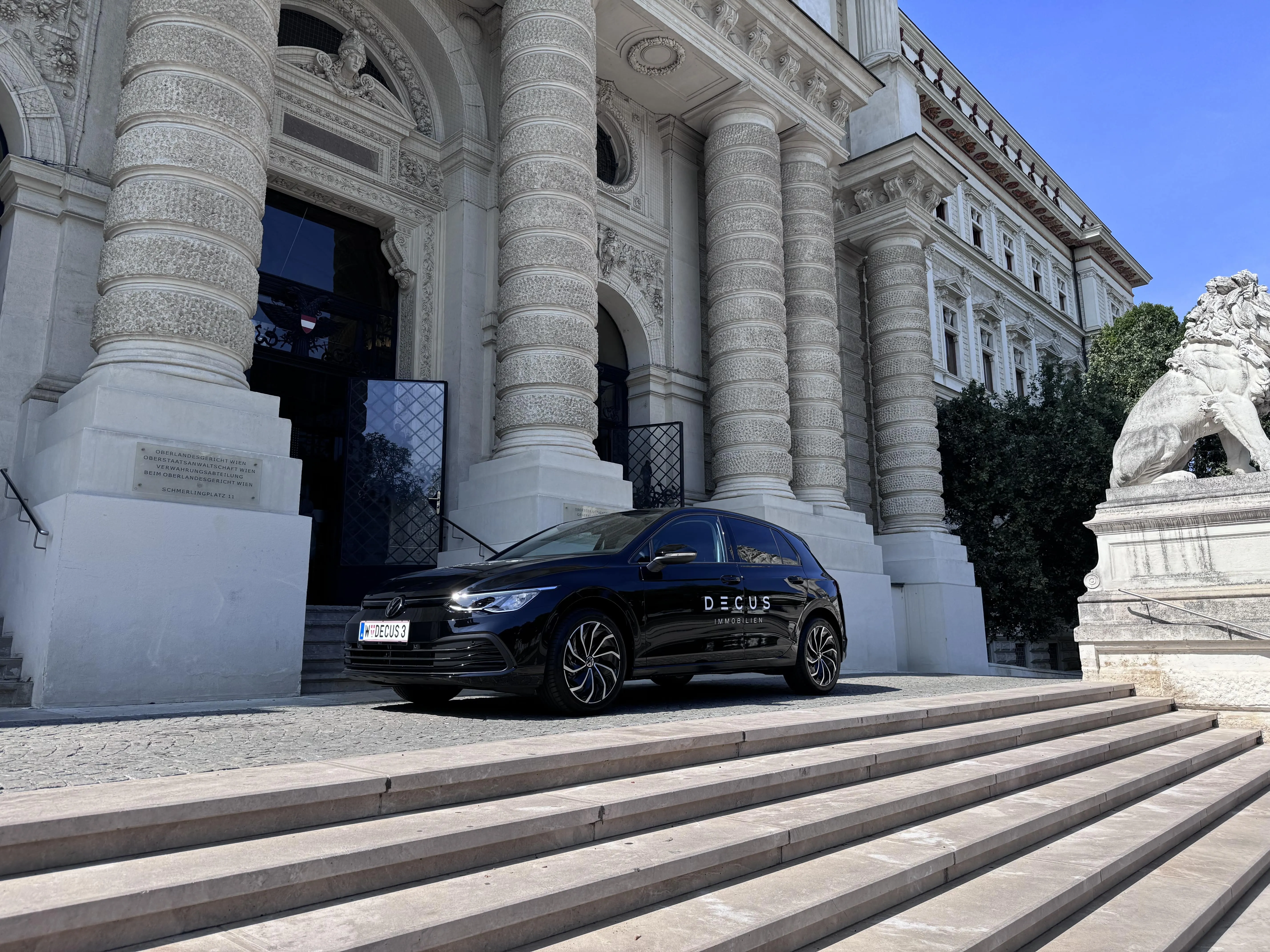 Black Volkswagen car with DECUS Immobilien branding parked in front of ornate stone columns and a lion statue at a grand historic building.