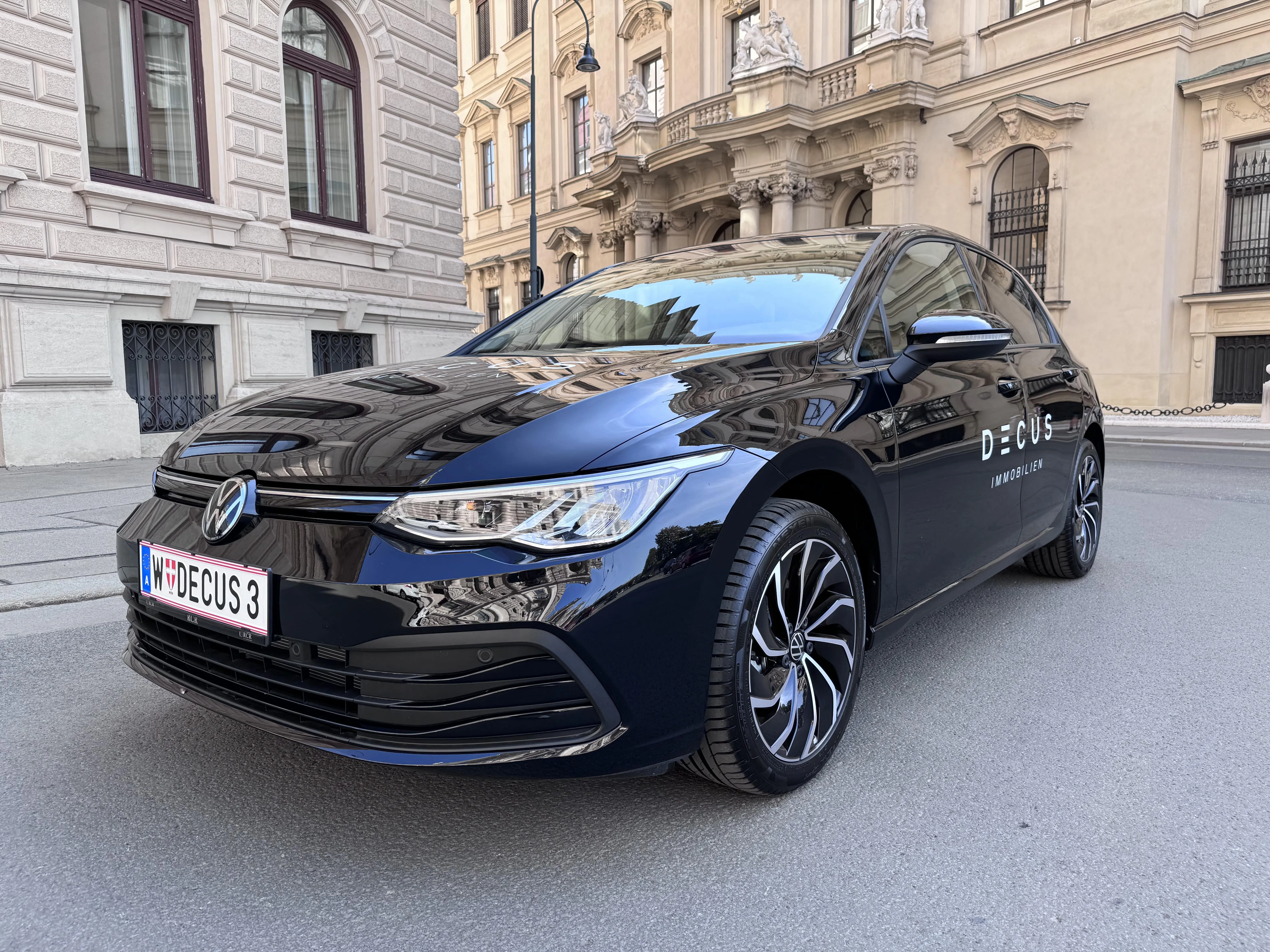 Black Volkswagen car with DECUS Immobilien branding parked on a city street with historic buildings reflected on its surface.