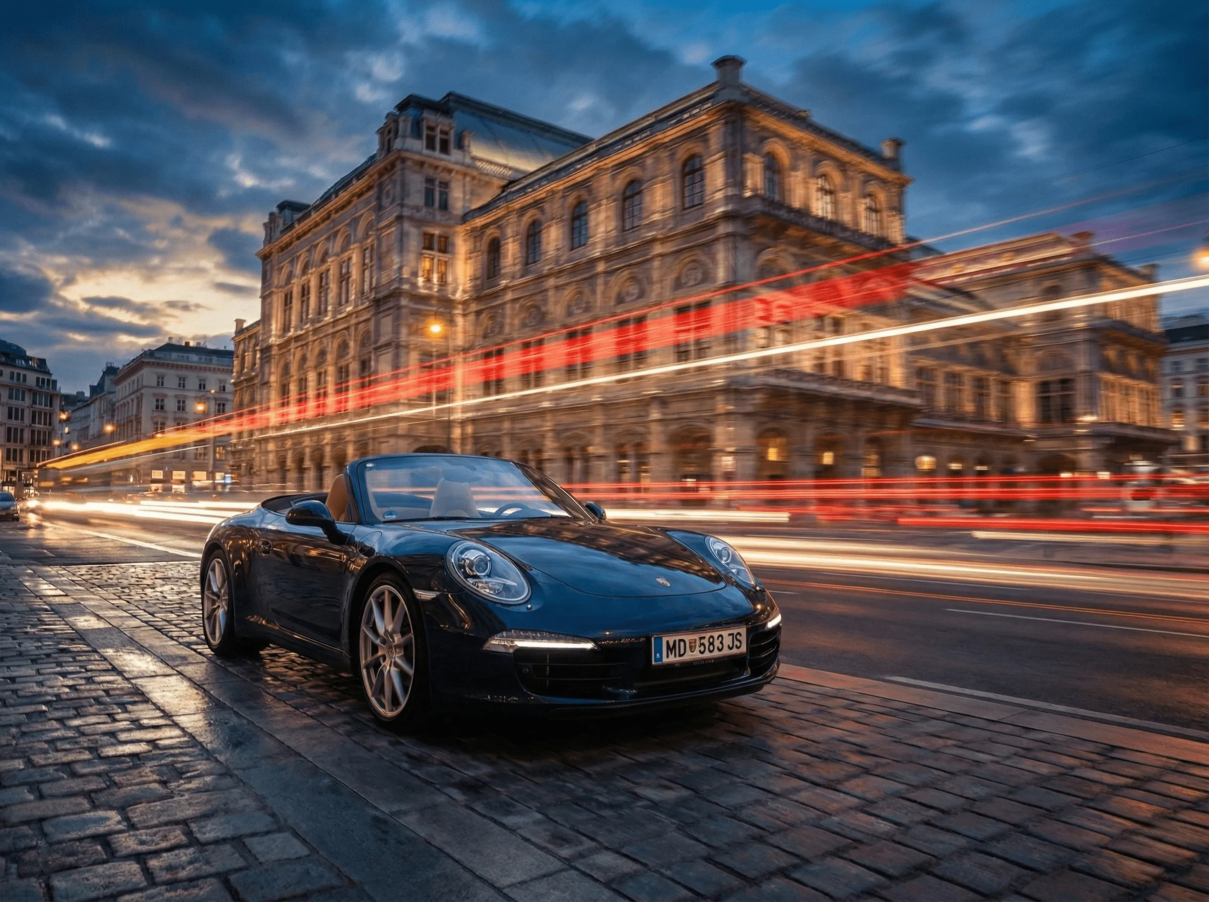 Black Porsche convertible parked on cobblestone street at dusk with historic illuminated building and streaks of car lights in background.