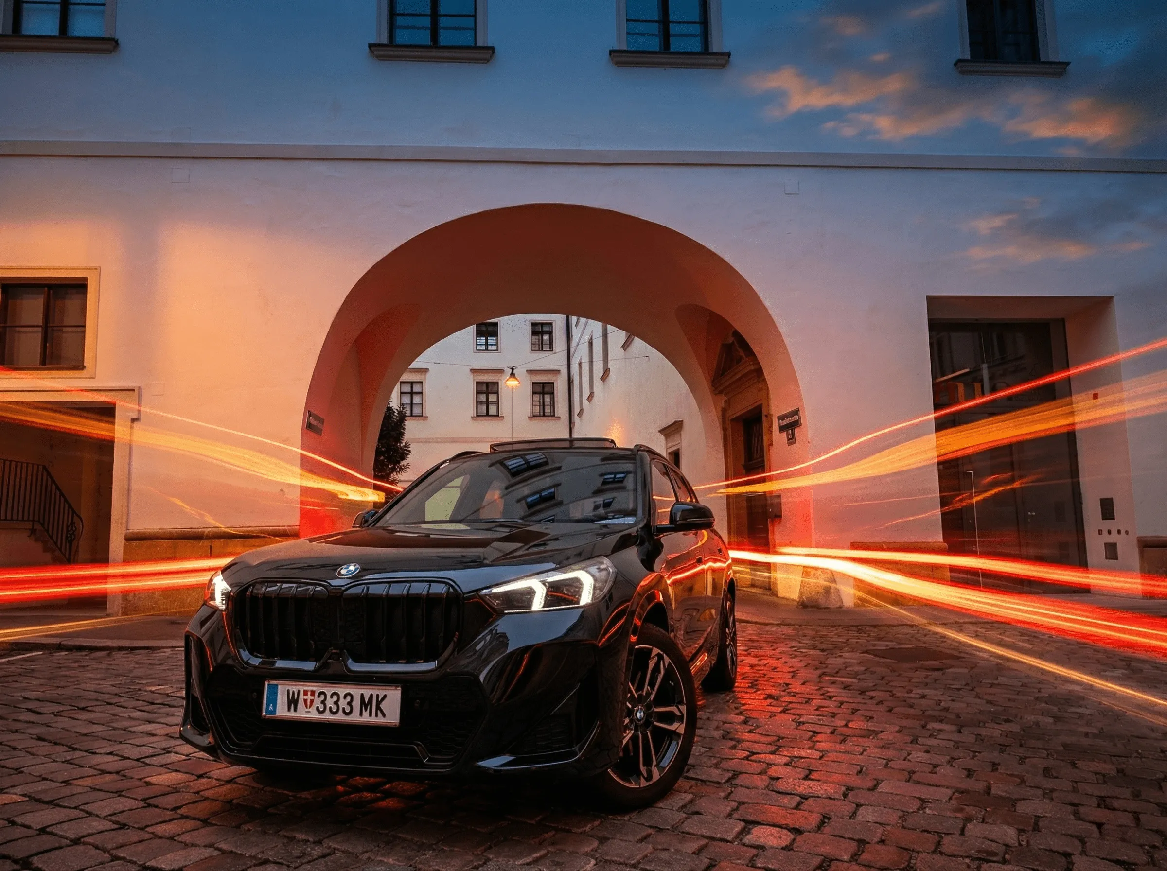 Black BMW car with headlights on parked on cobblestone street under an archway at dusk with streaks of orange light trails behind it.