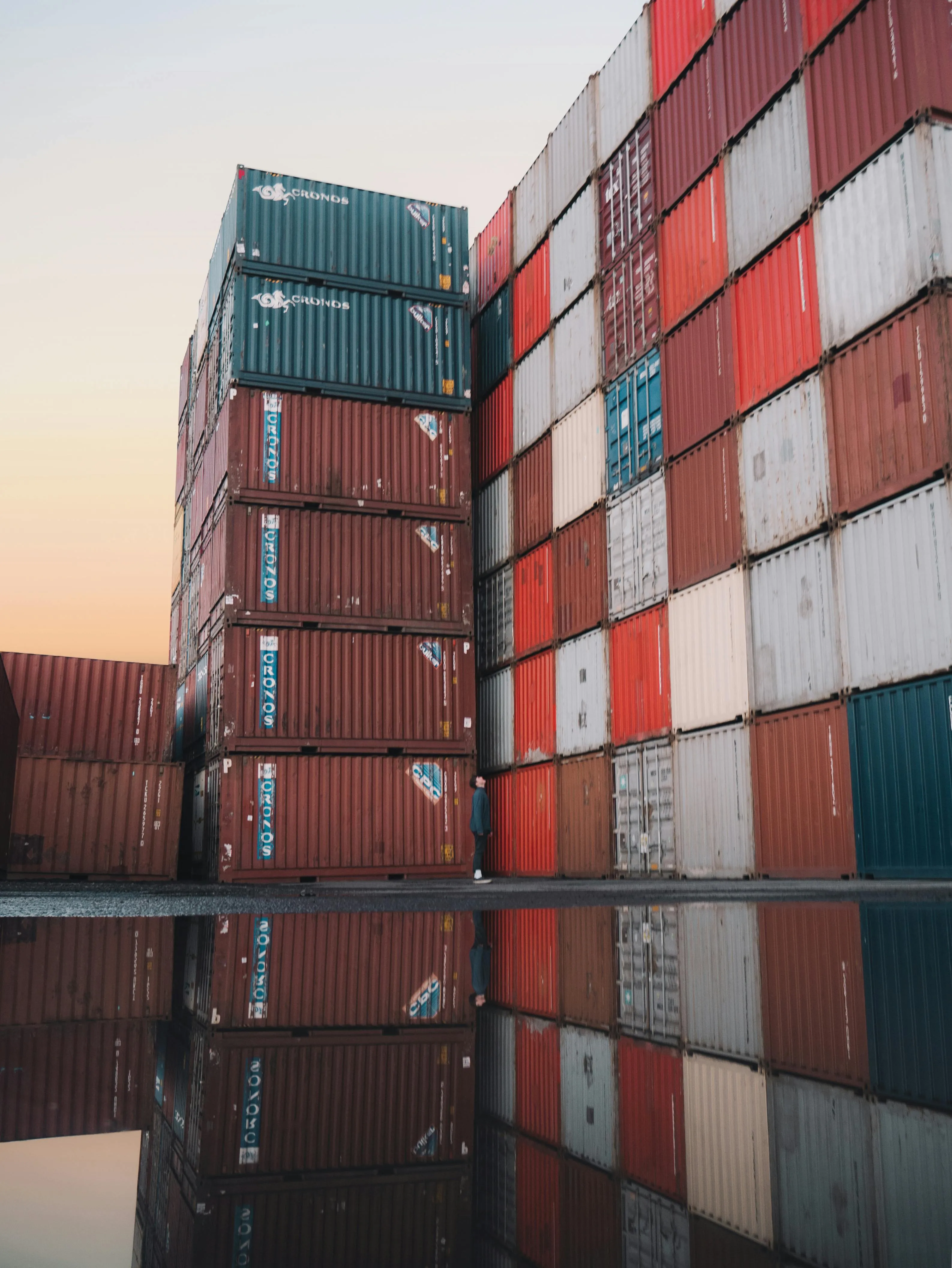 Person standing between large stacked shipping containers with their reflection visible in a water puddle.