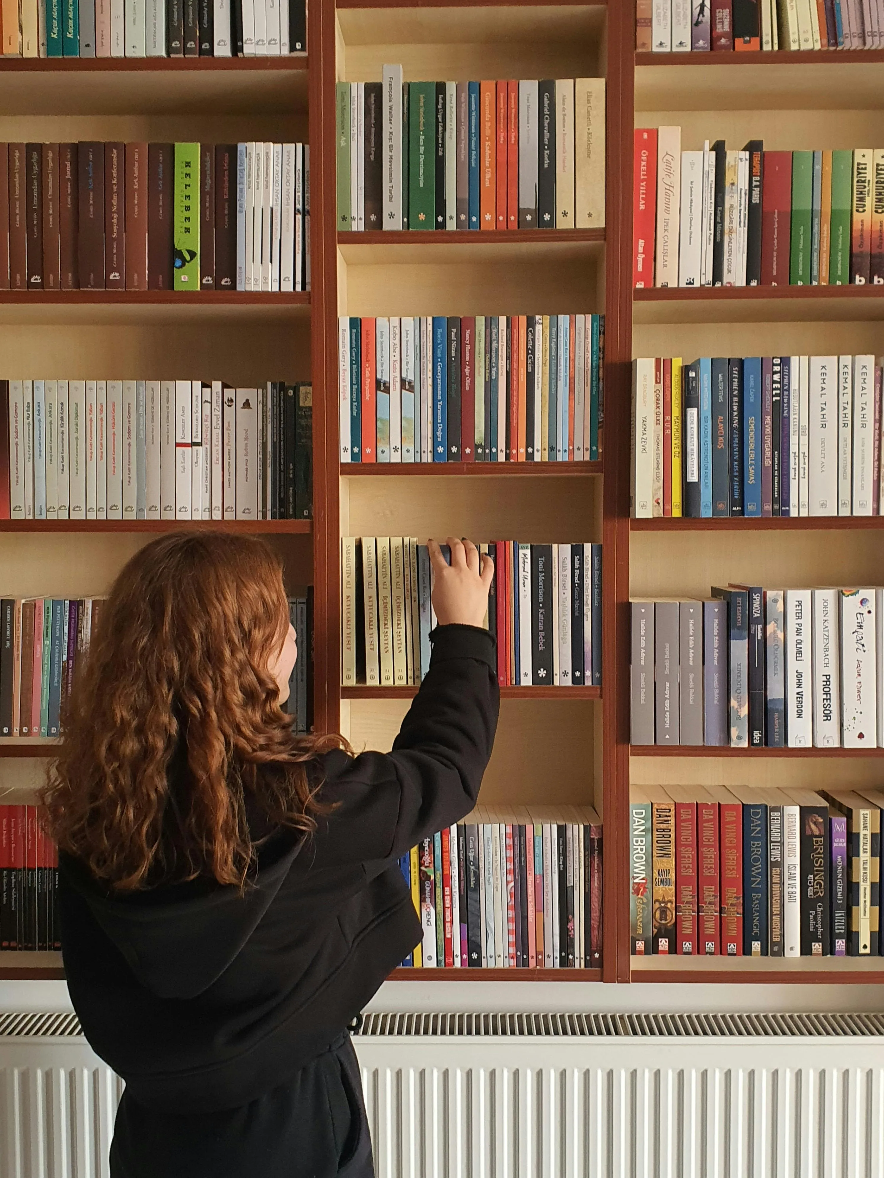 Person with curly red hair wearing a black hoodie reaching for a book on a wooden bookshelf filled with various books.
