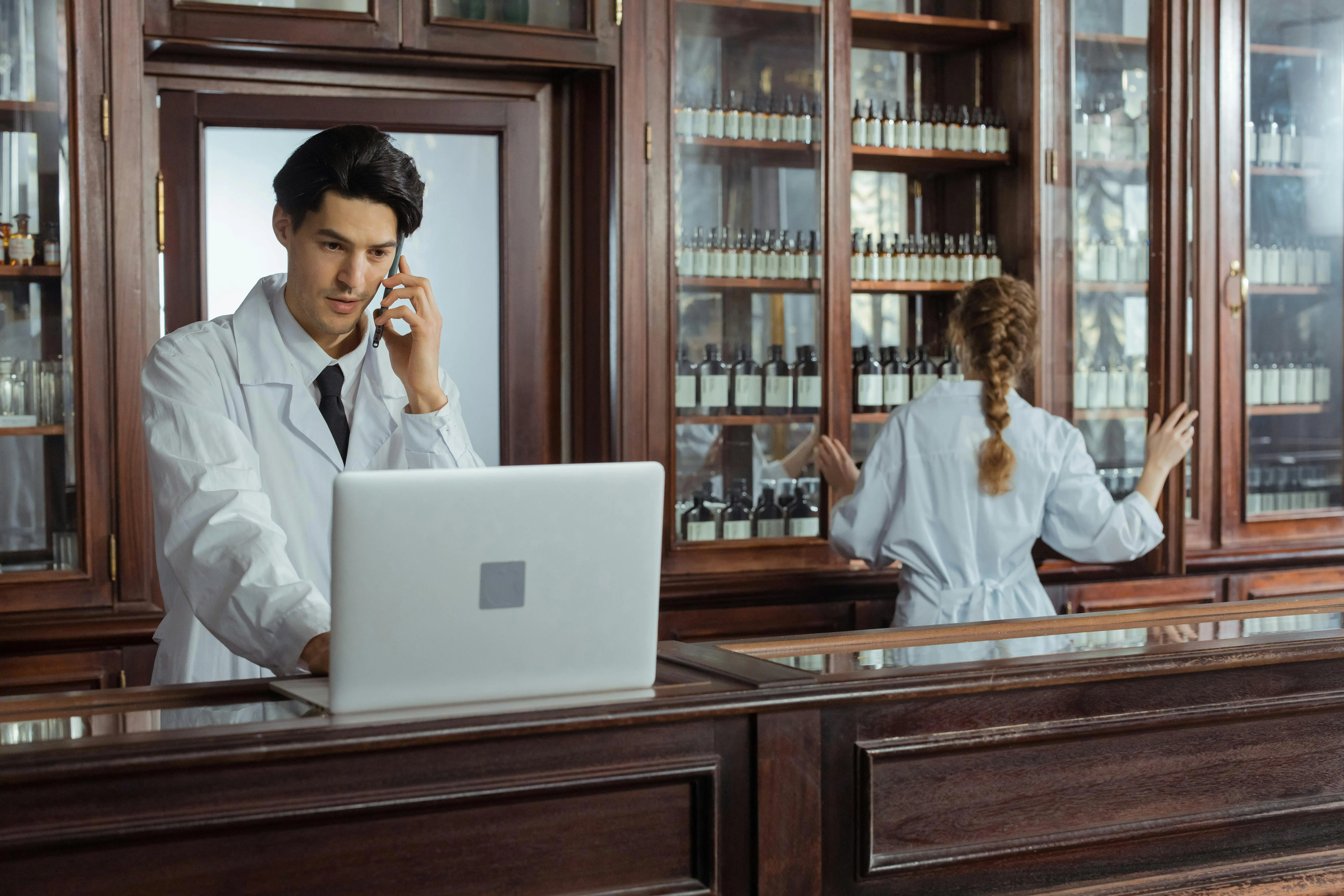 Two pharmacists in white coats working in a vintage-style pharmacy, one talking on the phone with a laptop in front of him, and the other organizing bottles in a cabinet.
