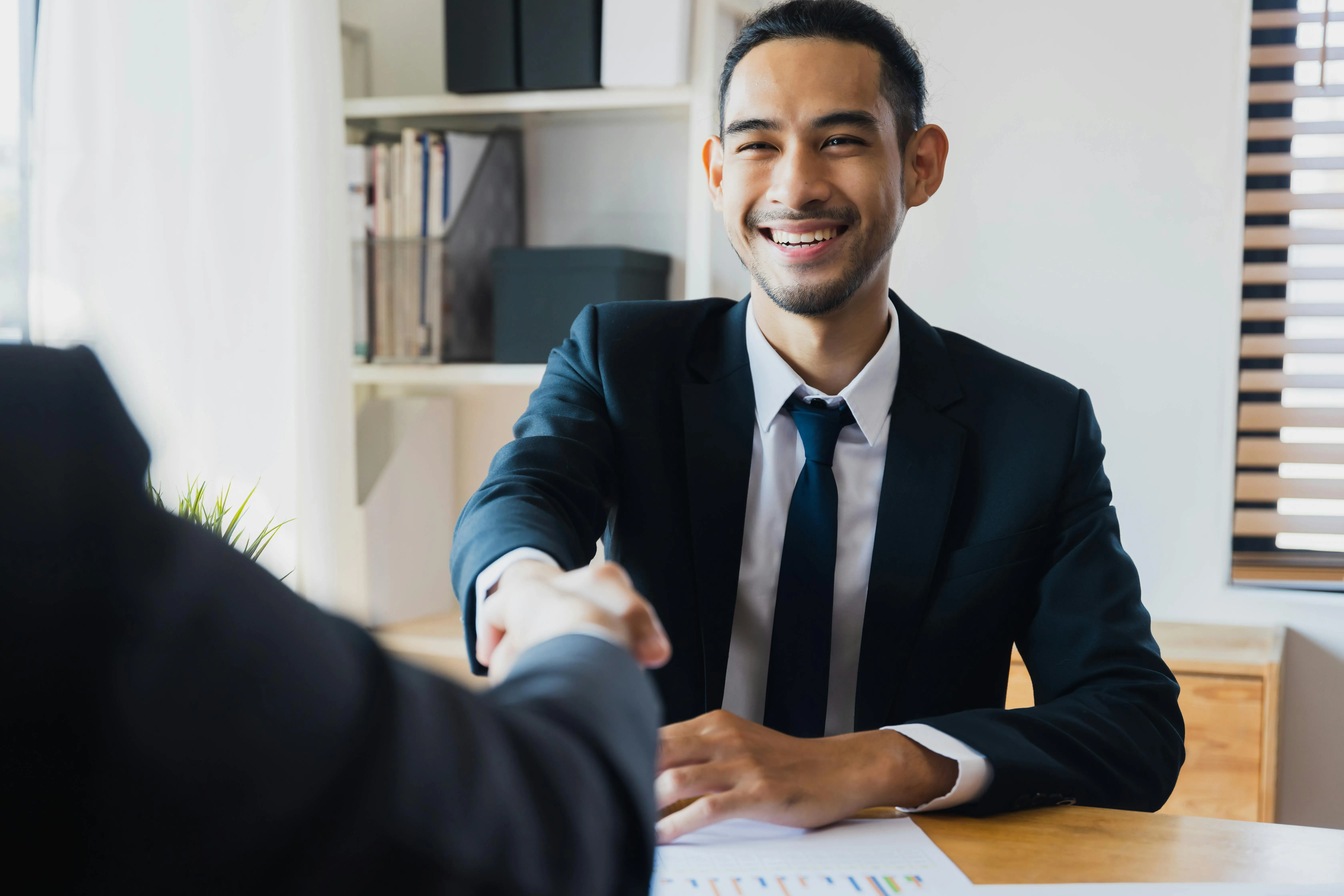 Smiling man in a suit shaking hands across a desk during a professional meeting.