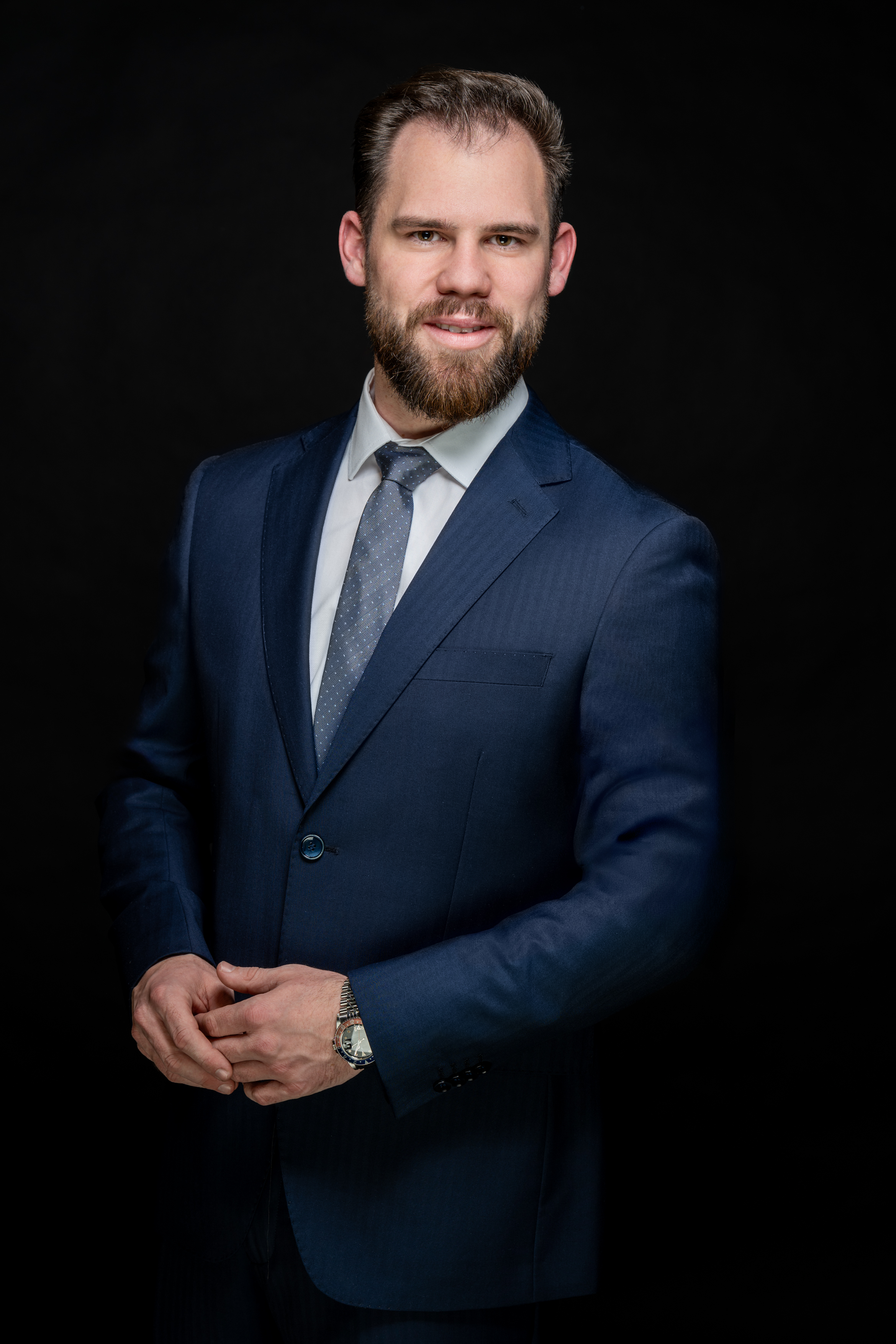 Smiling bearded man in a navy blue suit and patterned tie posing against a black background.