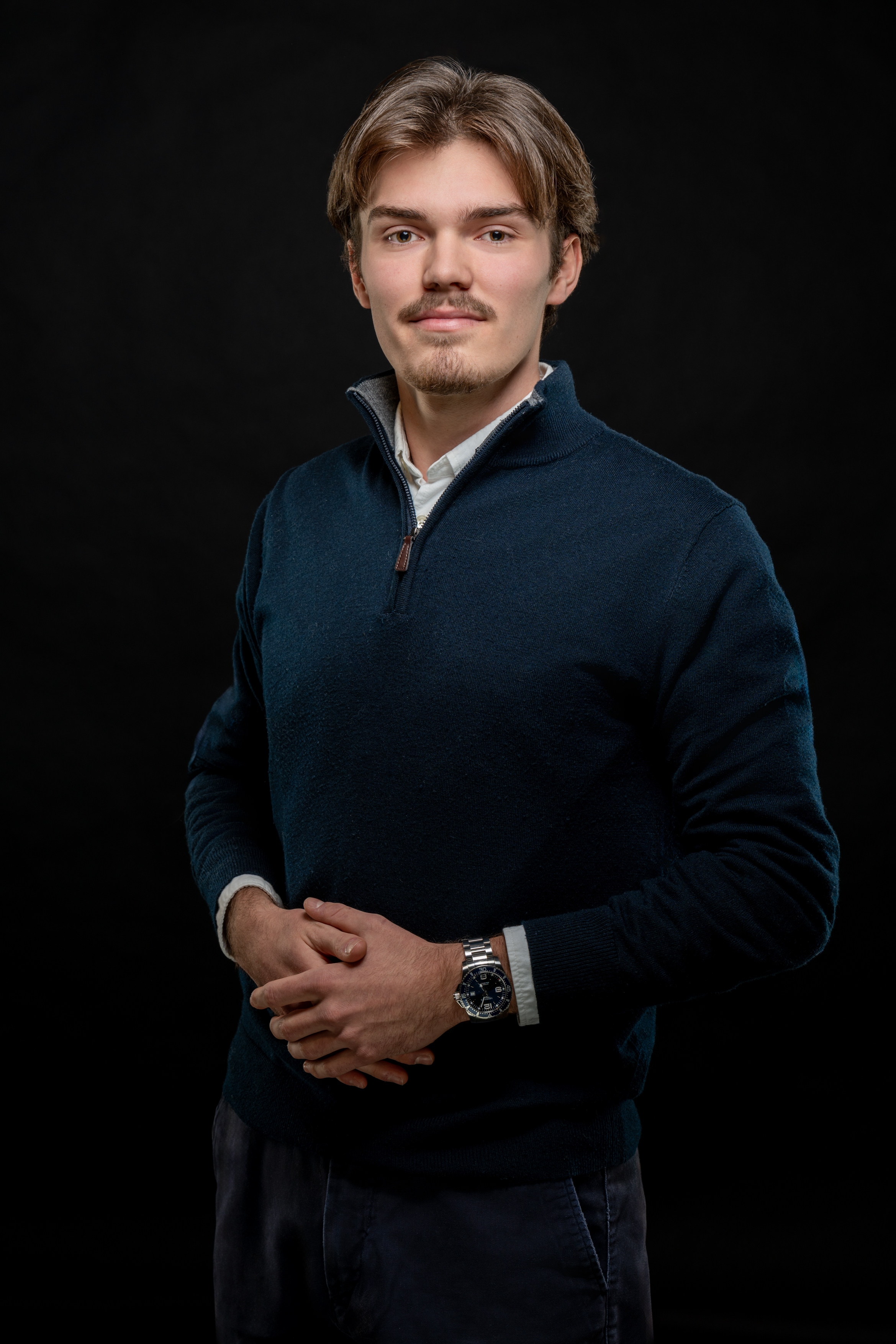 Young man with light brown hair and a mustache, wearing a dark blue quarter-zip sweater and watch, standing against a black background.