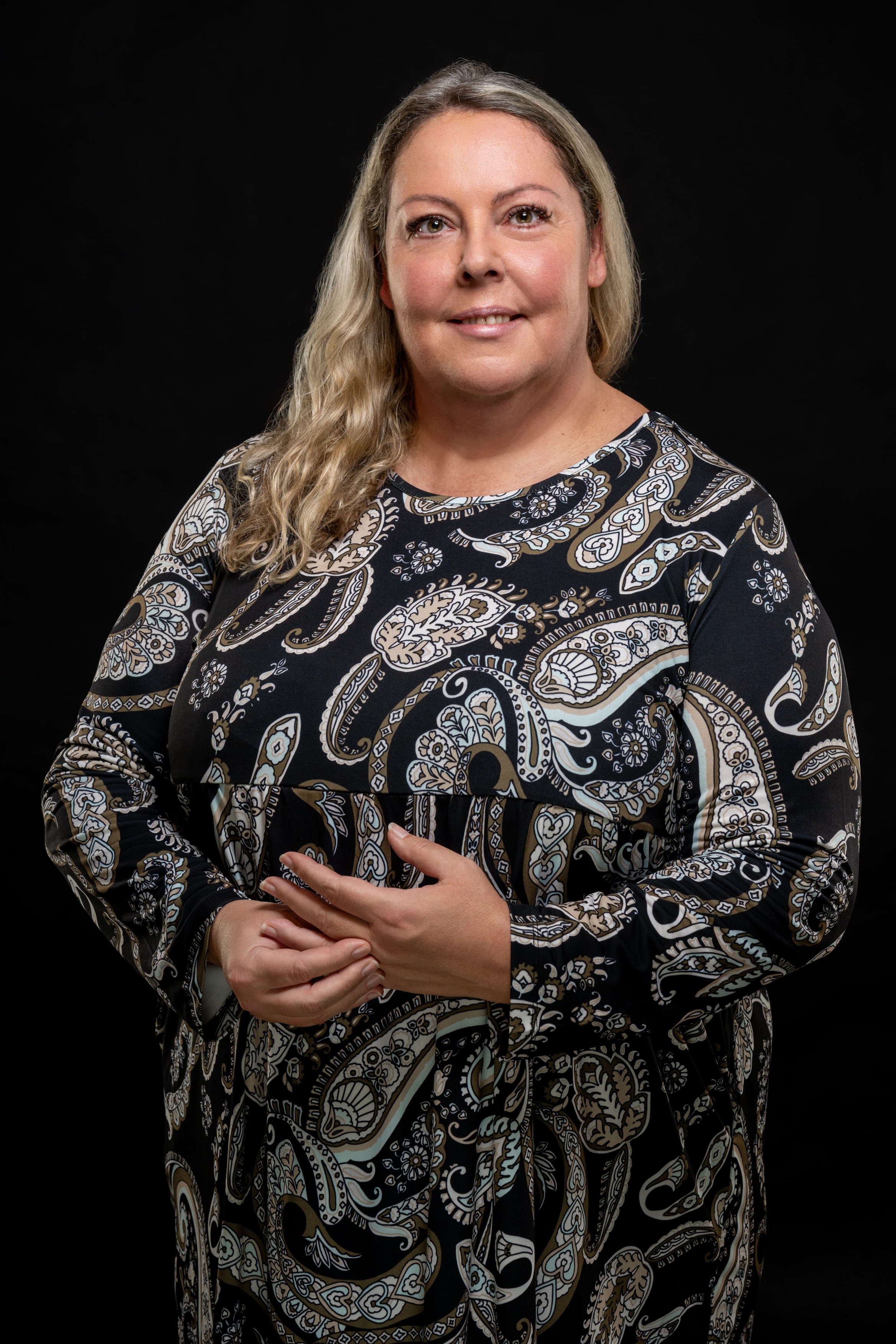 Woman with long blonde hair wearing a black dress with beige and white paisley patterns, standing against a black background.