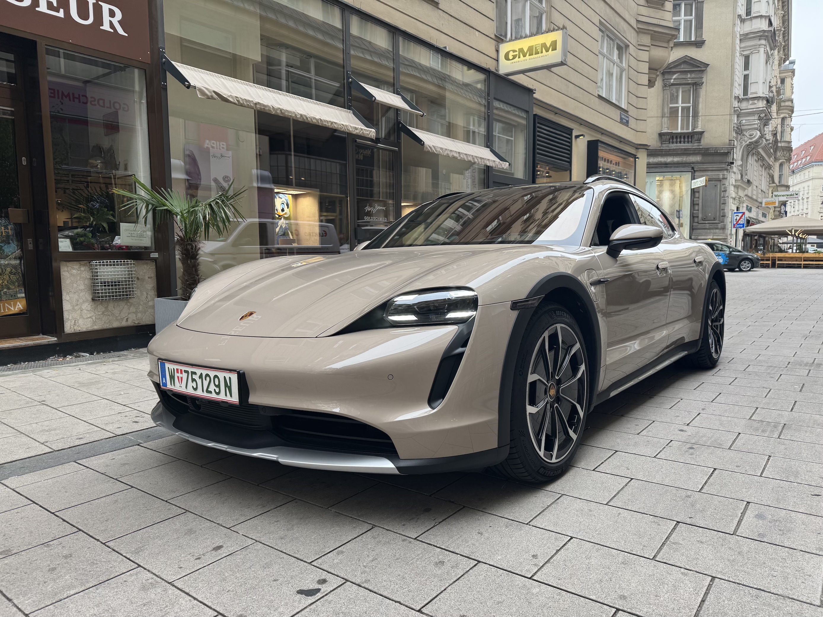 Beige Porsche Taycan parked on a city street near buildings and shops.