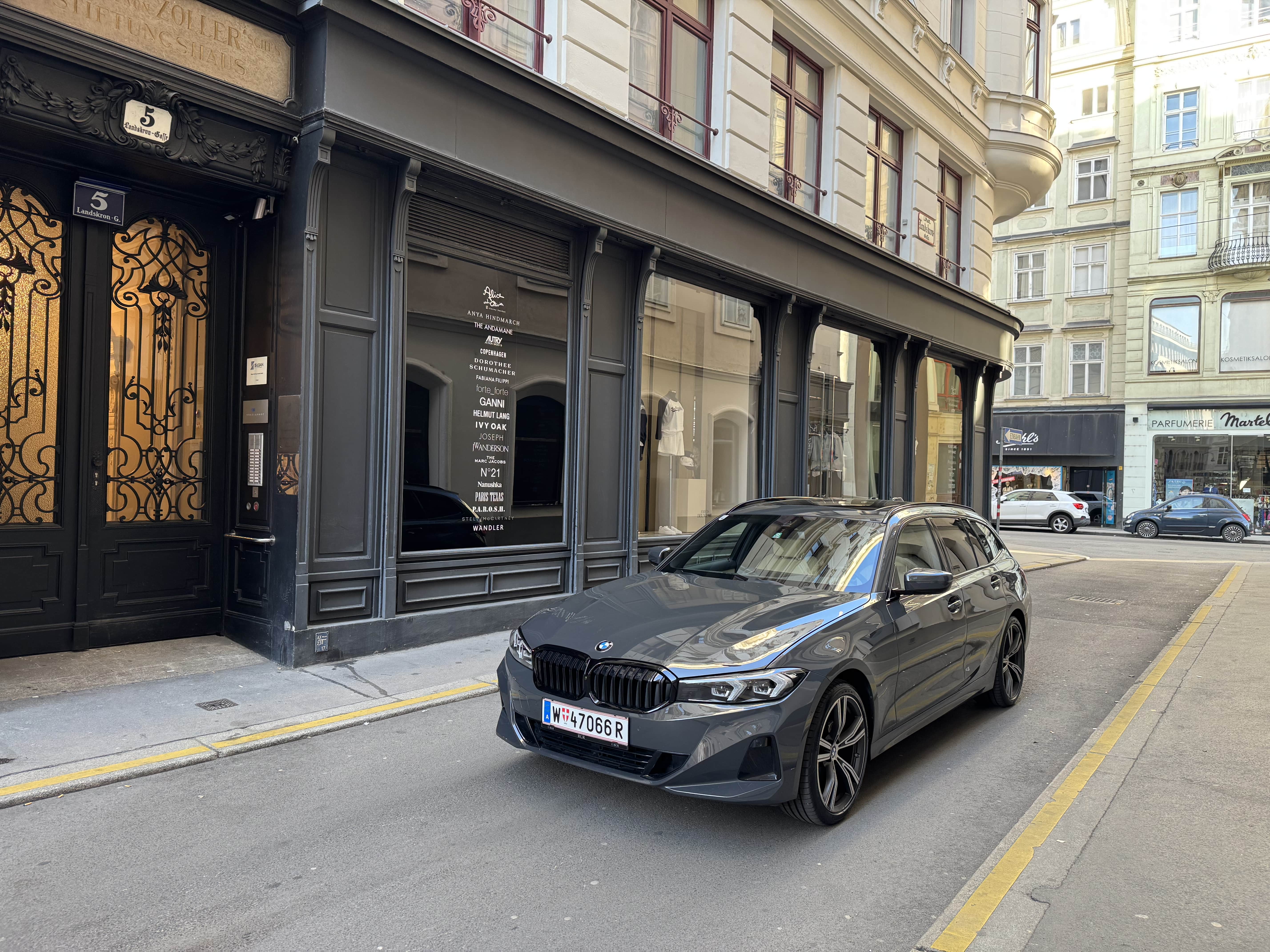Gray BMW car parked on a narrow street in front of a building with large windows and ornate black doors.