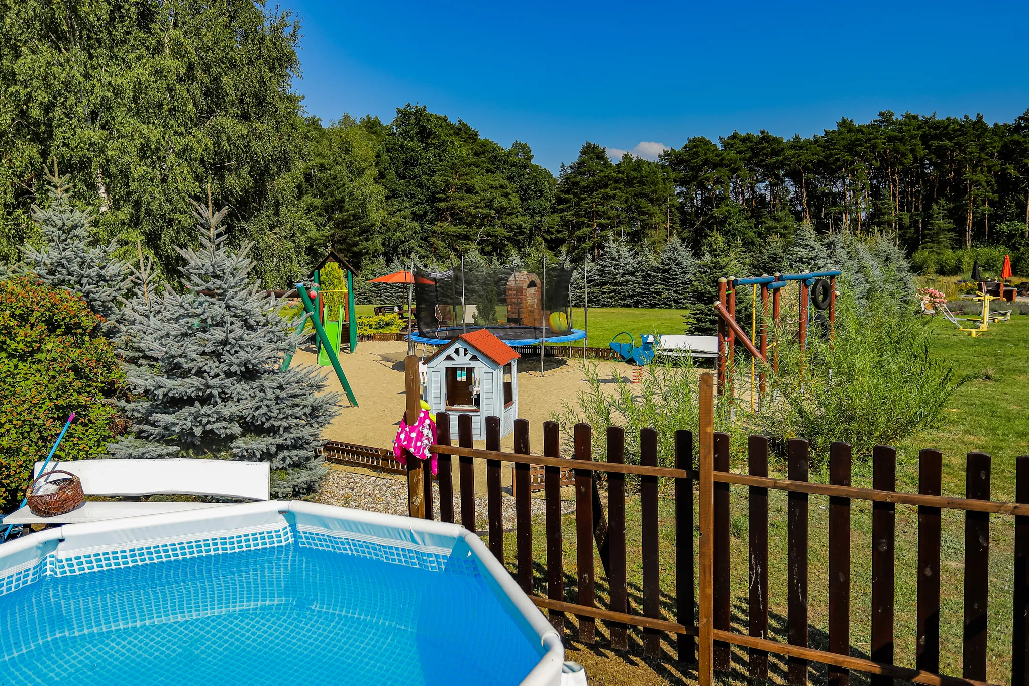 Basen ogrodowy na pierwszym planie z placem zabaw i huśtawkami otoczonymi zielenią i drzewami w tle.