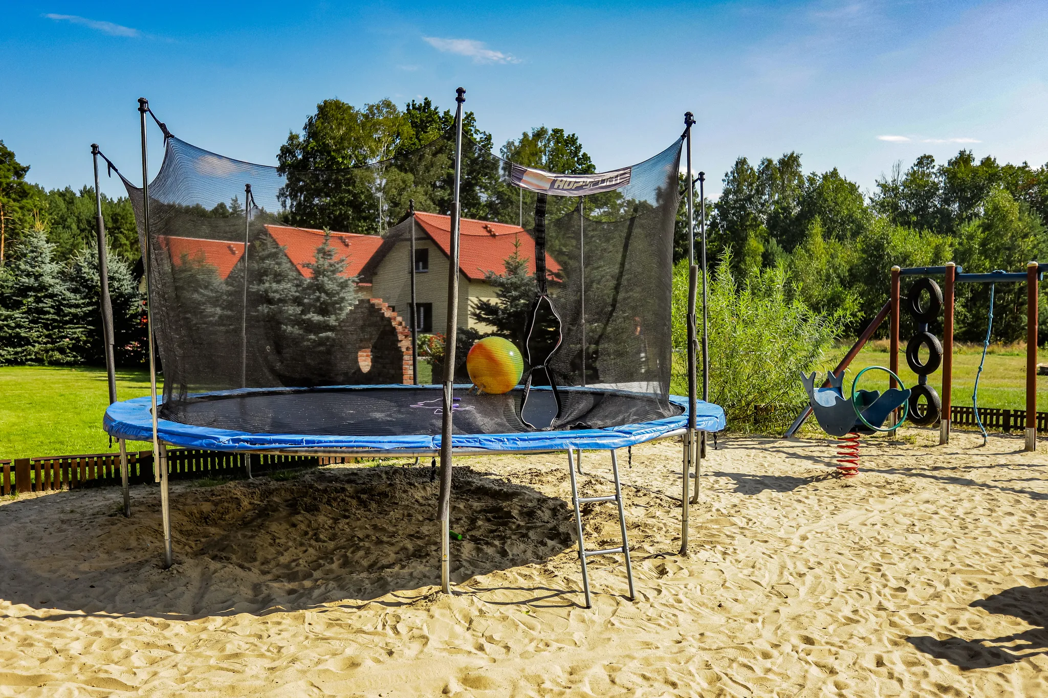 Plac zabaw z trampoliną na piasku, dużą piłką i huśtawkami na tle zielonych drzew i domu z czerwonym dachem.