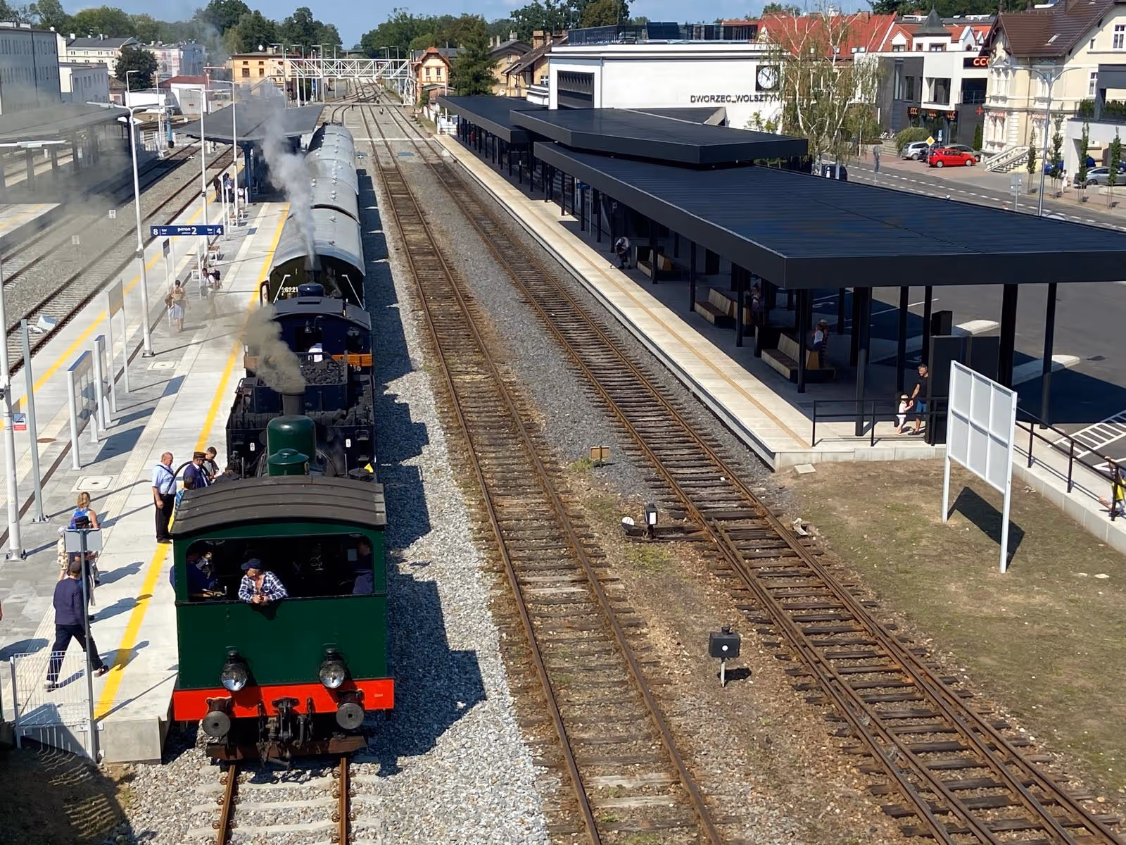 Stara czarna i zielona lokomotywa parowa z wagonami stoi na peronie dworca kolejowego w słoneczny dzień.