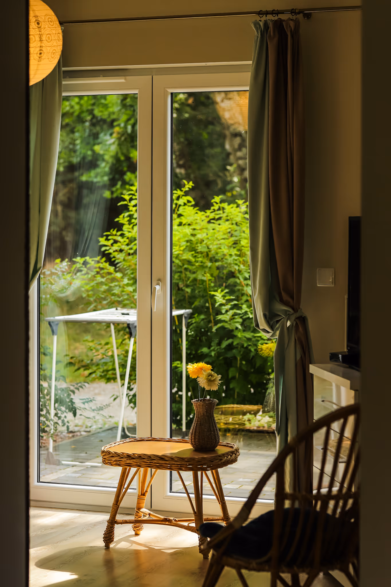 Mały stolik z wikliny ze szklaną wazonem i żółtymi kwiatami przy dużych drzwiach tarasowych z widokiem na zielony ogród.