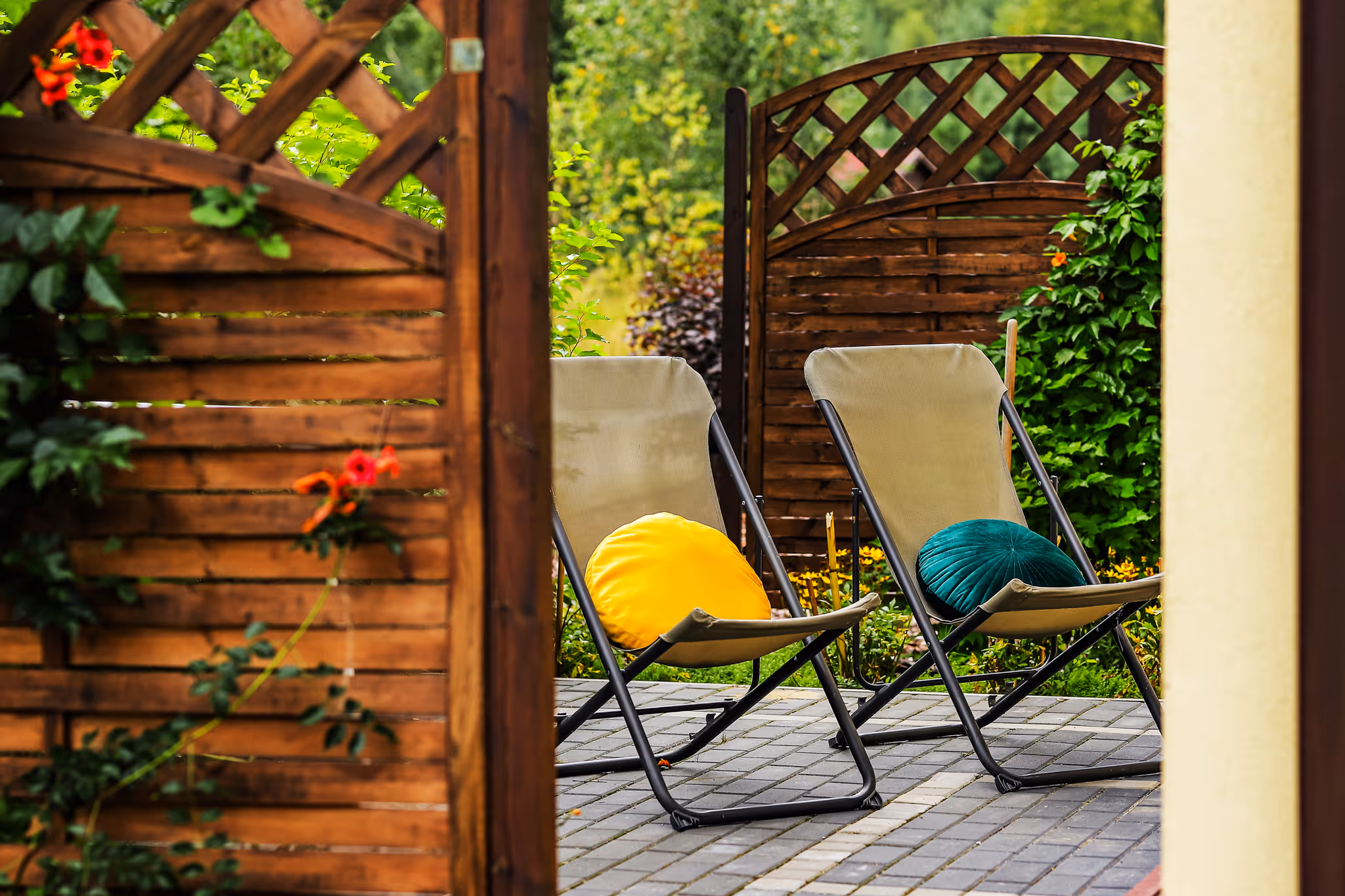 Dwa składane krzesła na patio z żółtą i zieloną okrągłą poduszką, otoczone drewnianym płotem i zielenią.