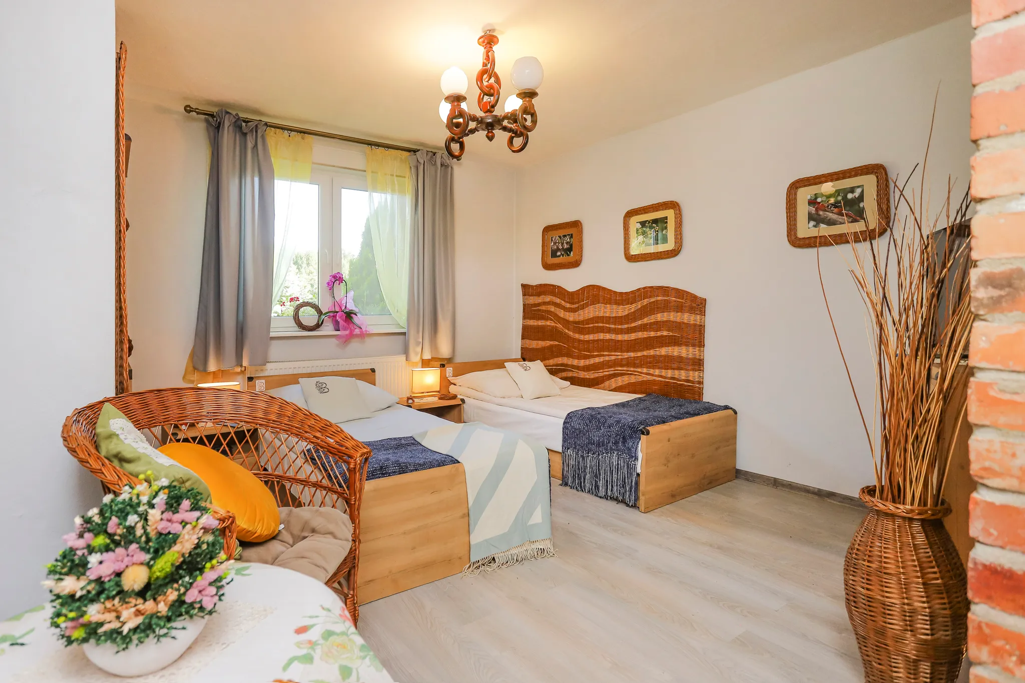 Przytulna sypialnia z łóżkiem, żółtymi zasłonami, telewizorem na komodzie oraz wiklinowymi meblami przy ścianie z czerwonej cegły.