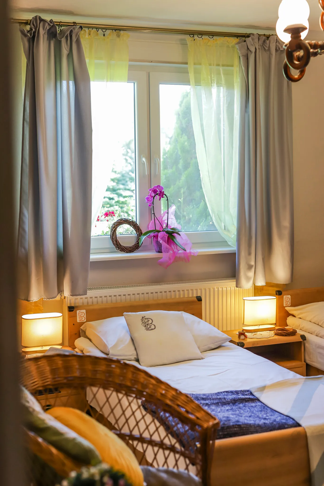 Przytulna sypialnia z łóżkiem, żółtymi zasłonami, telewizorem na komodzie oraz wiklinowymi meblami przy ścianie z czerwonej cegły.